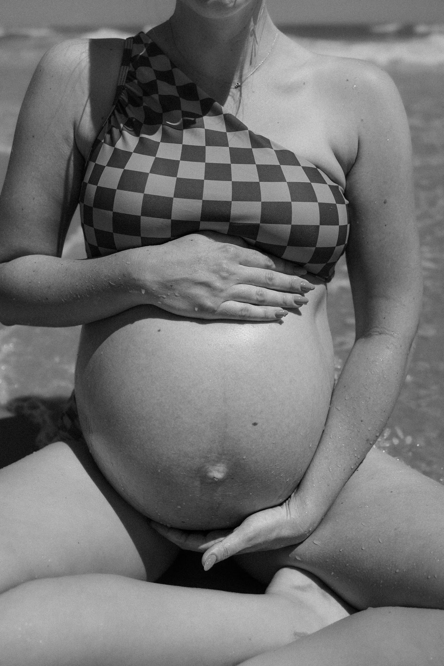 Black and white photo of a pregnant woman in a checkered bikini top cradling her baby bump at the beach.