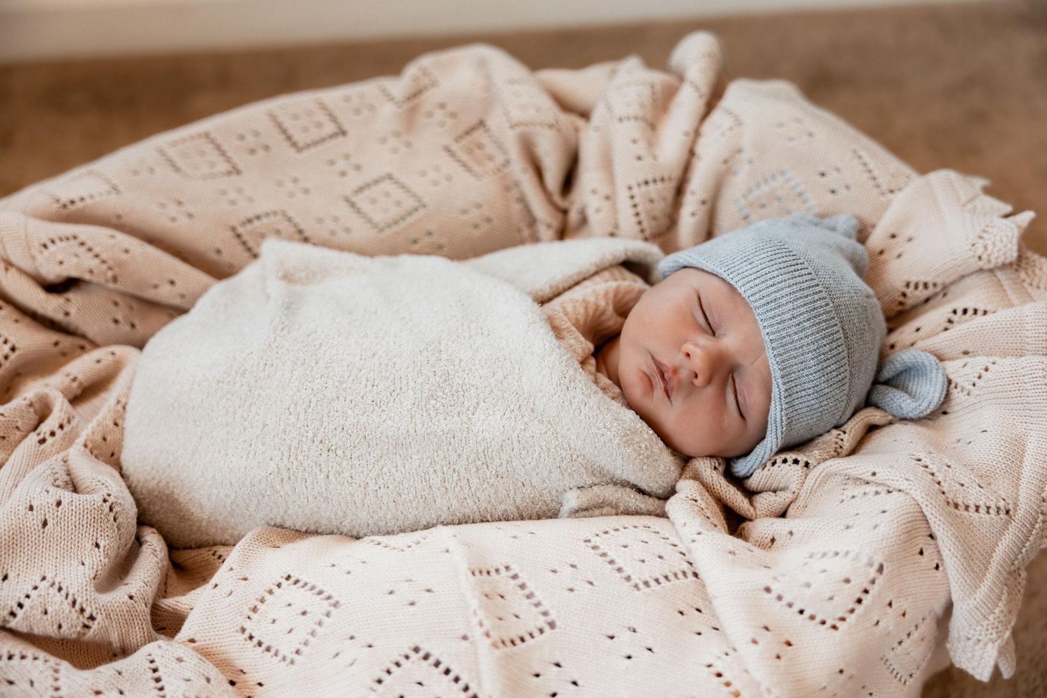Sleeping newborn baby swaddled in white knit wrap wearing blue hat resting in basket with soft pink blanket.