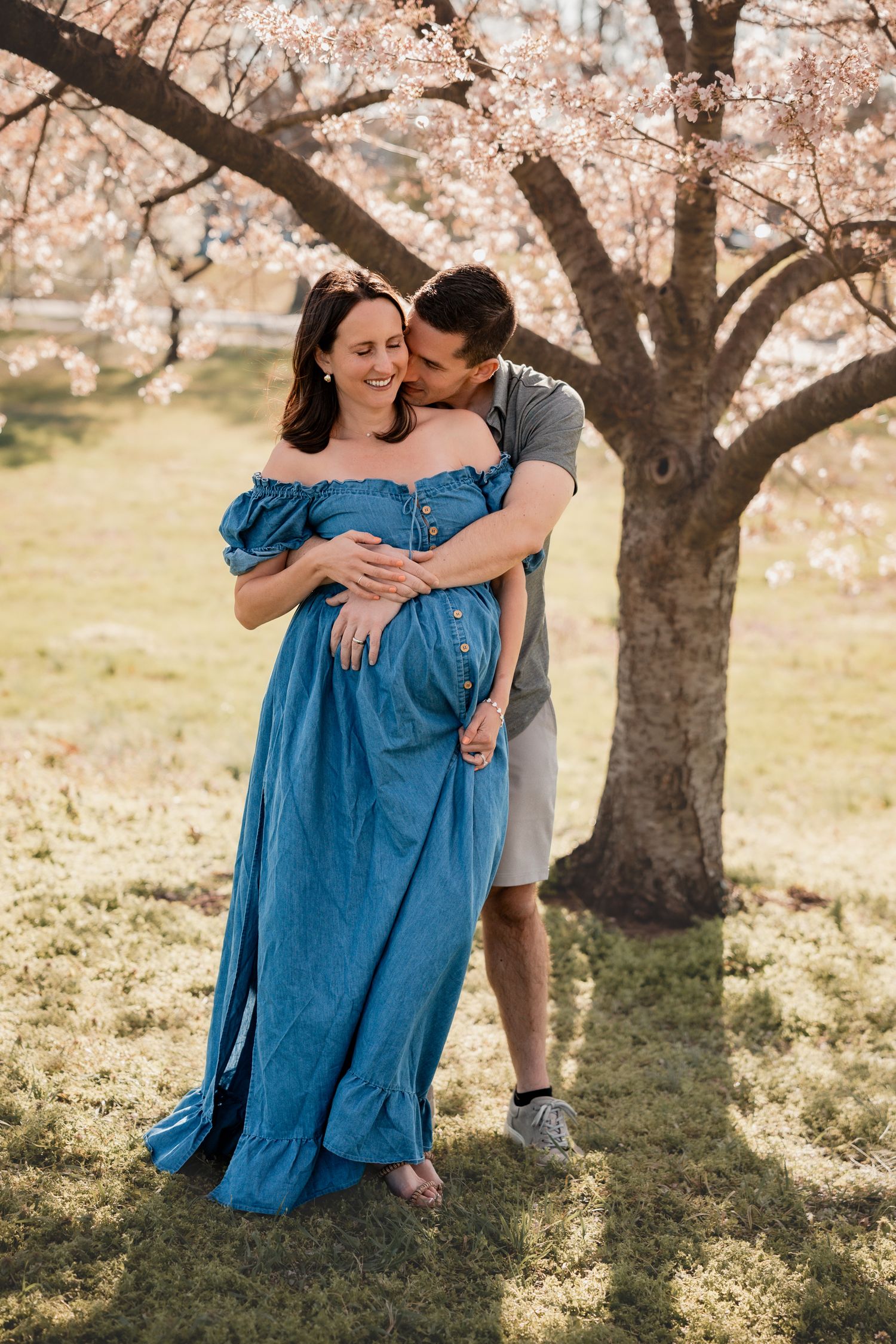 Couple sharing a tender moment under blooming cherry blossom tree, pregnant woman in blue off-shoulder dress.