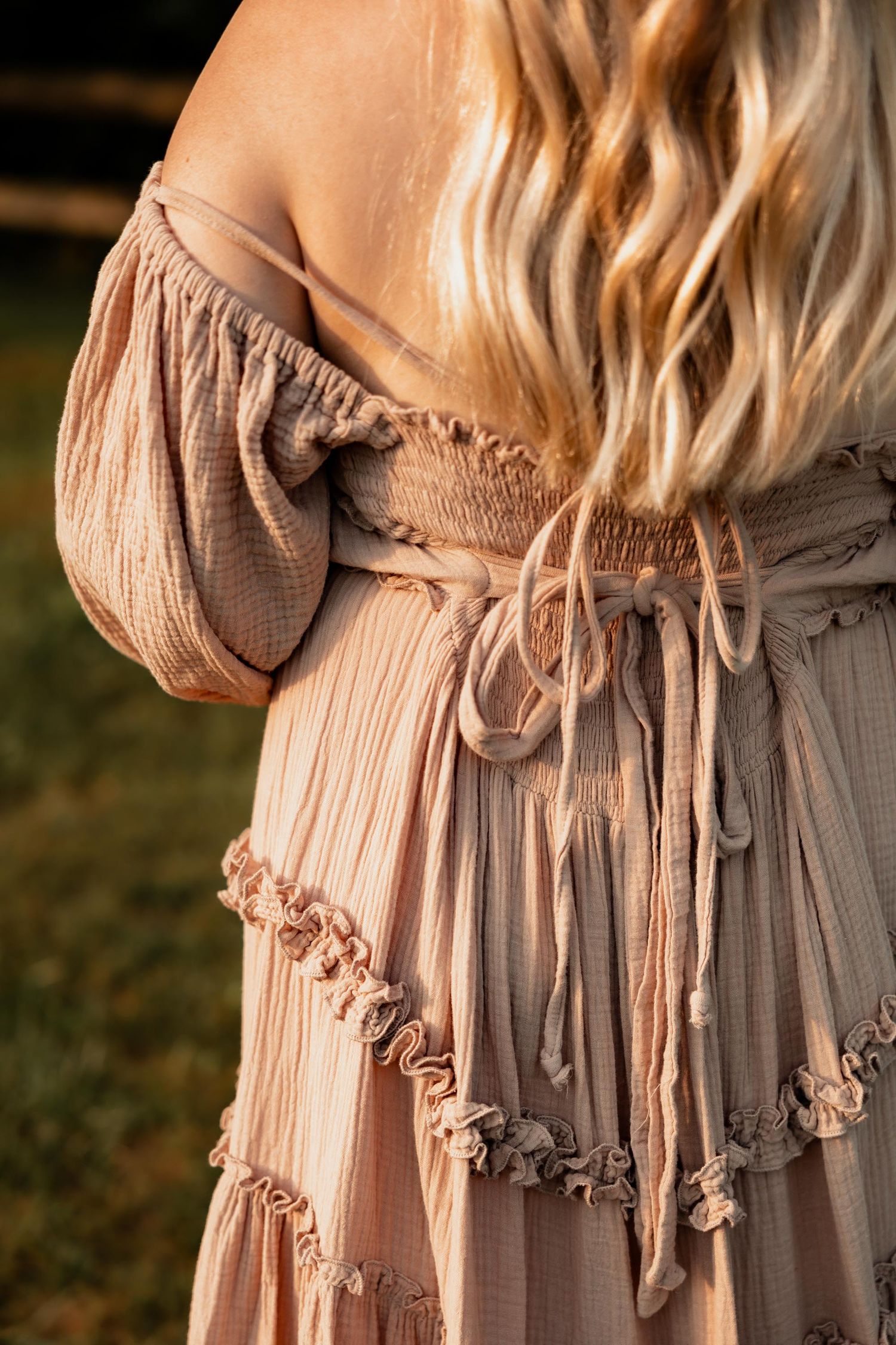 Blonde woman in blush pink off-shoulder boho dress with ruffled hem and tied waist sash, photographed outdoors.