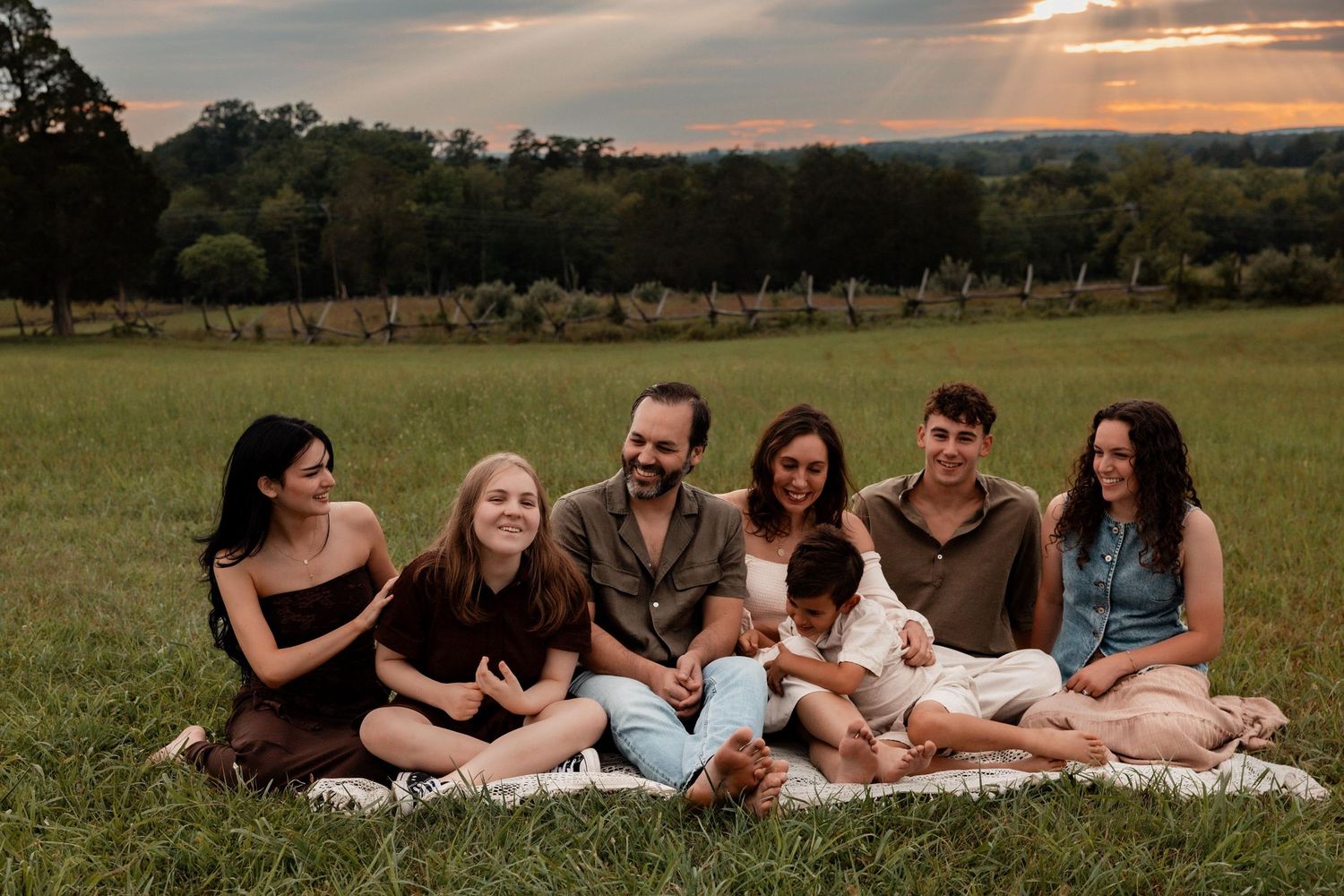 Family of seven sitting on a blanket in a green field at sunset, smiling together for a portrait.