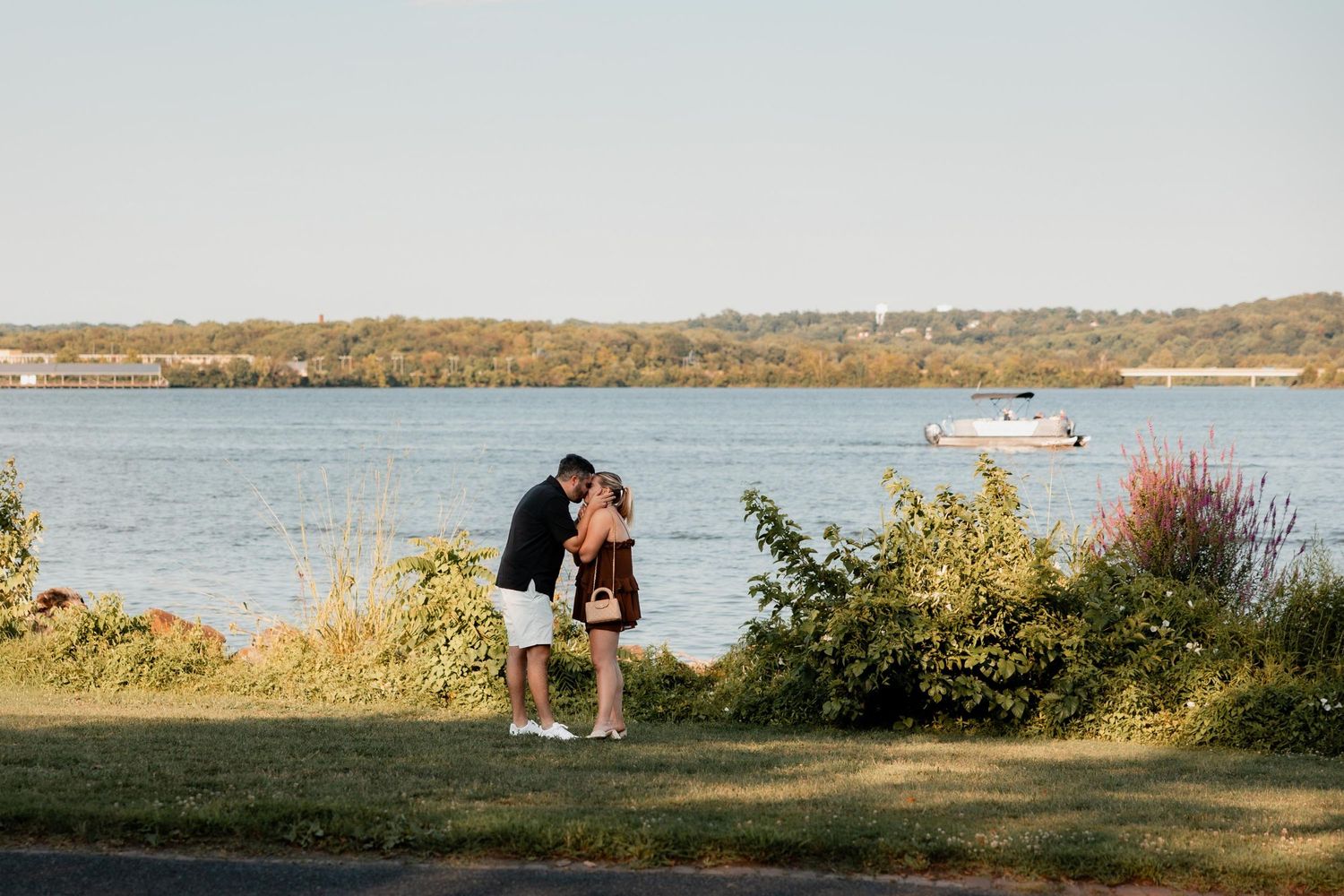 Couple sharing a romantic kiss by a scenic lakeside with lush greenery and a boat on the water in the background.