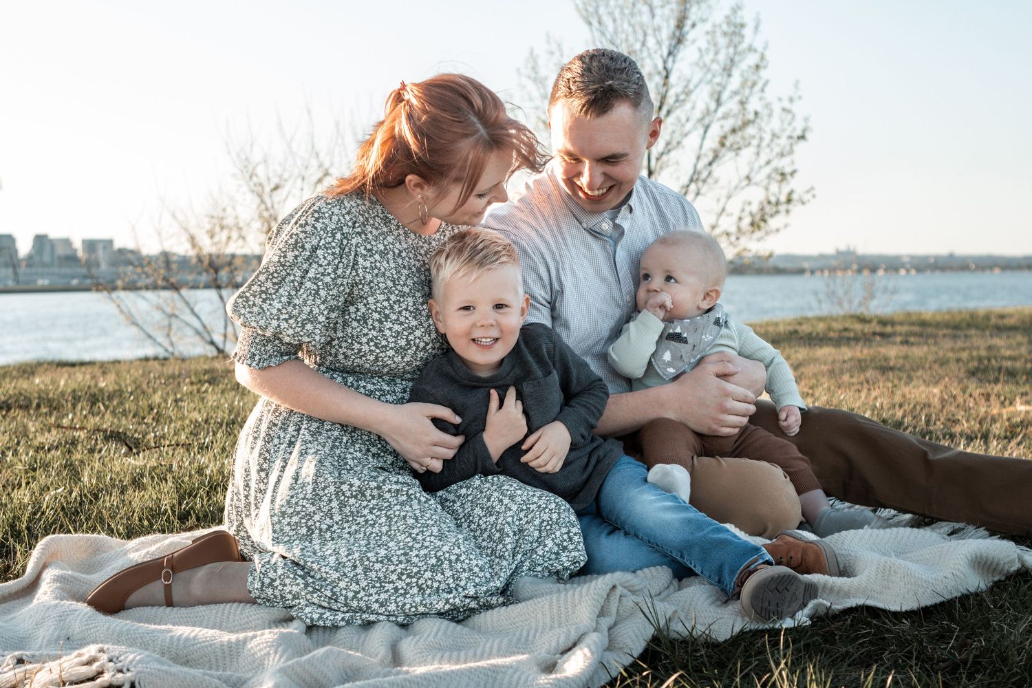 A loving family sits together on a blanket during a sunset picnic by the water on a grassy hillside.