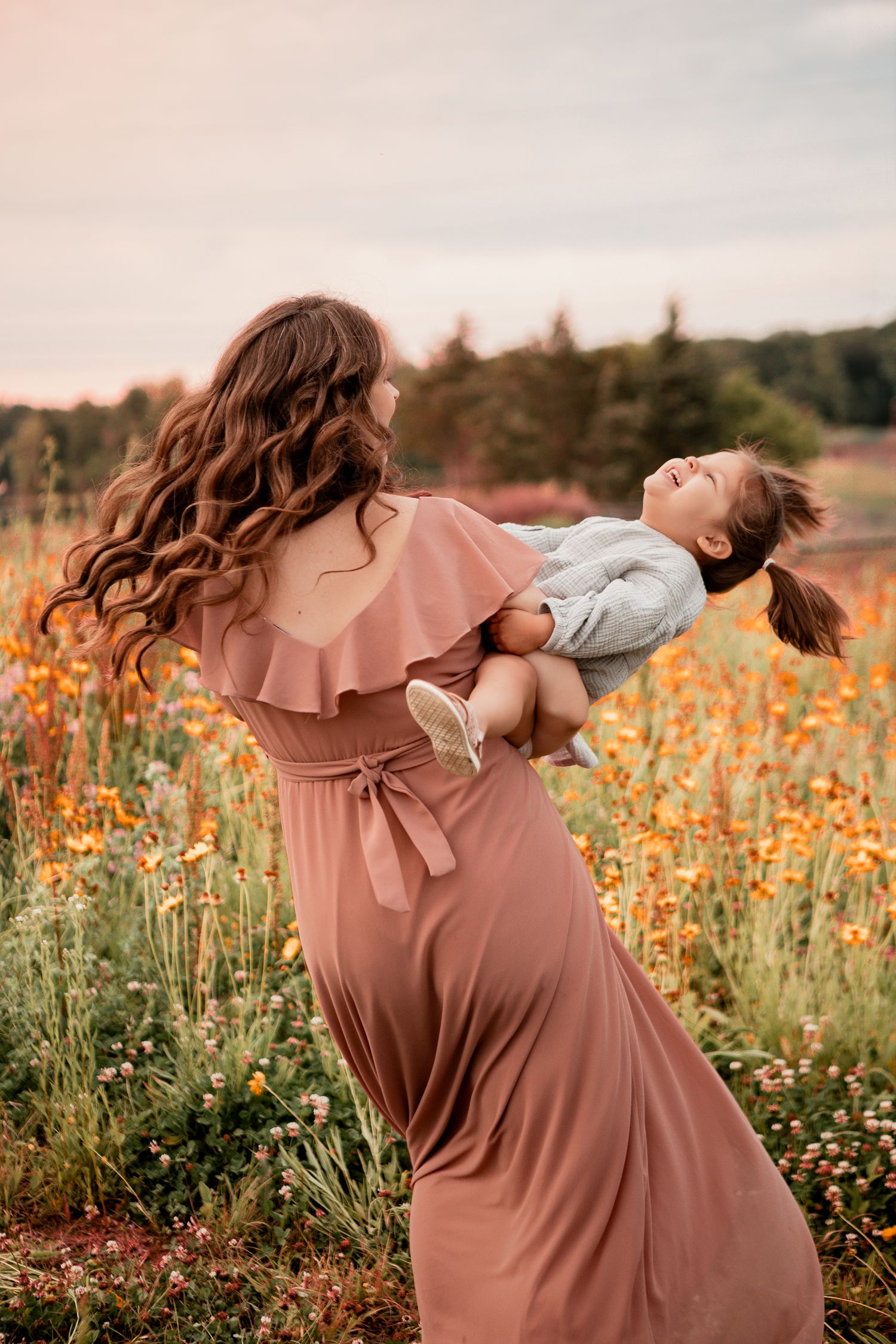 Two people share a joyful moment in a field of orange wildflowers at sunset, with one wearing a flowing dusty rose dress.