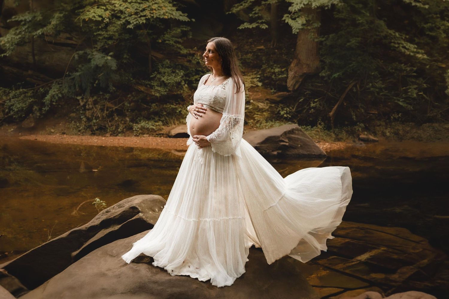 Pregnant woman in flowing white lace gown standing on rocks by a forest stream, cradling her baby bump.