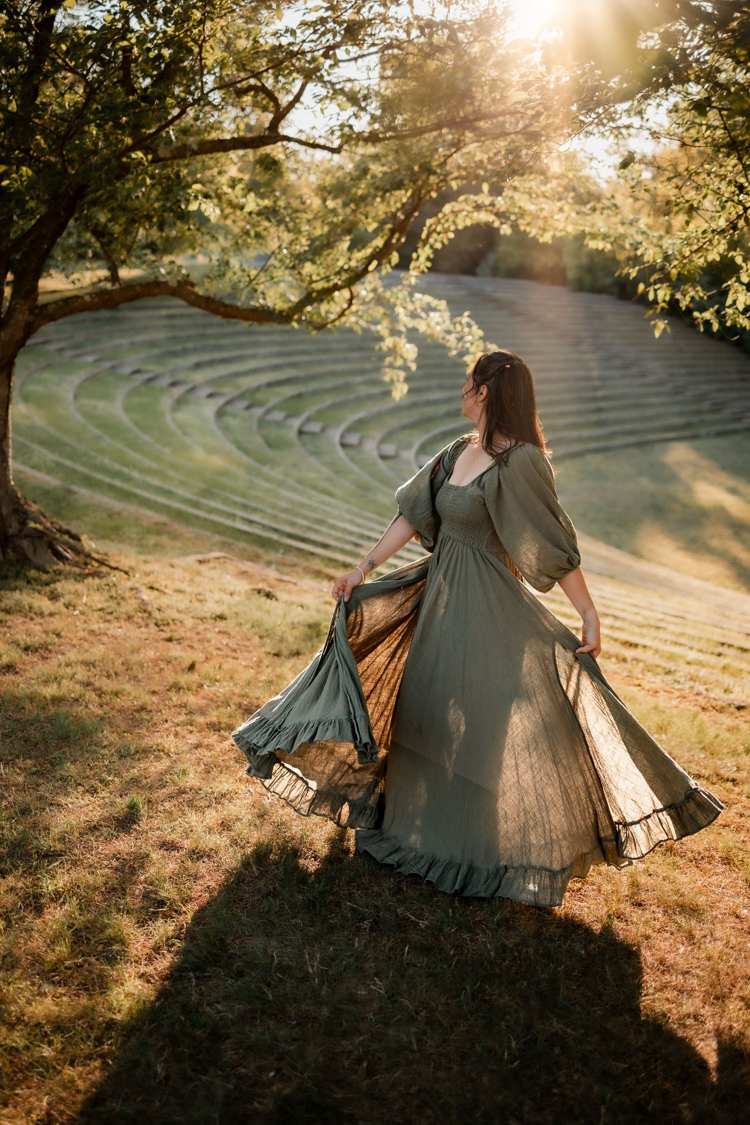 Woman in flowing sage green dress twirling at golden hour near terraced garden landscape.