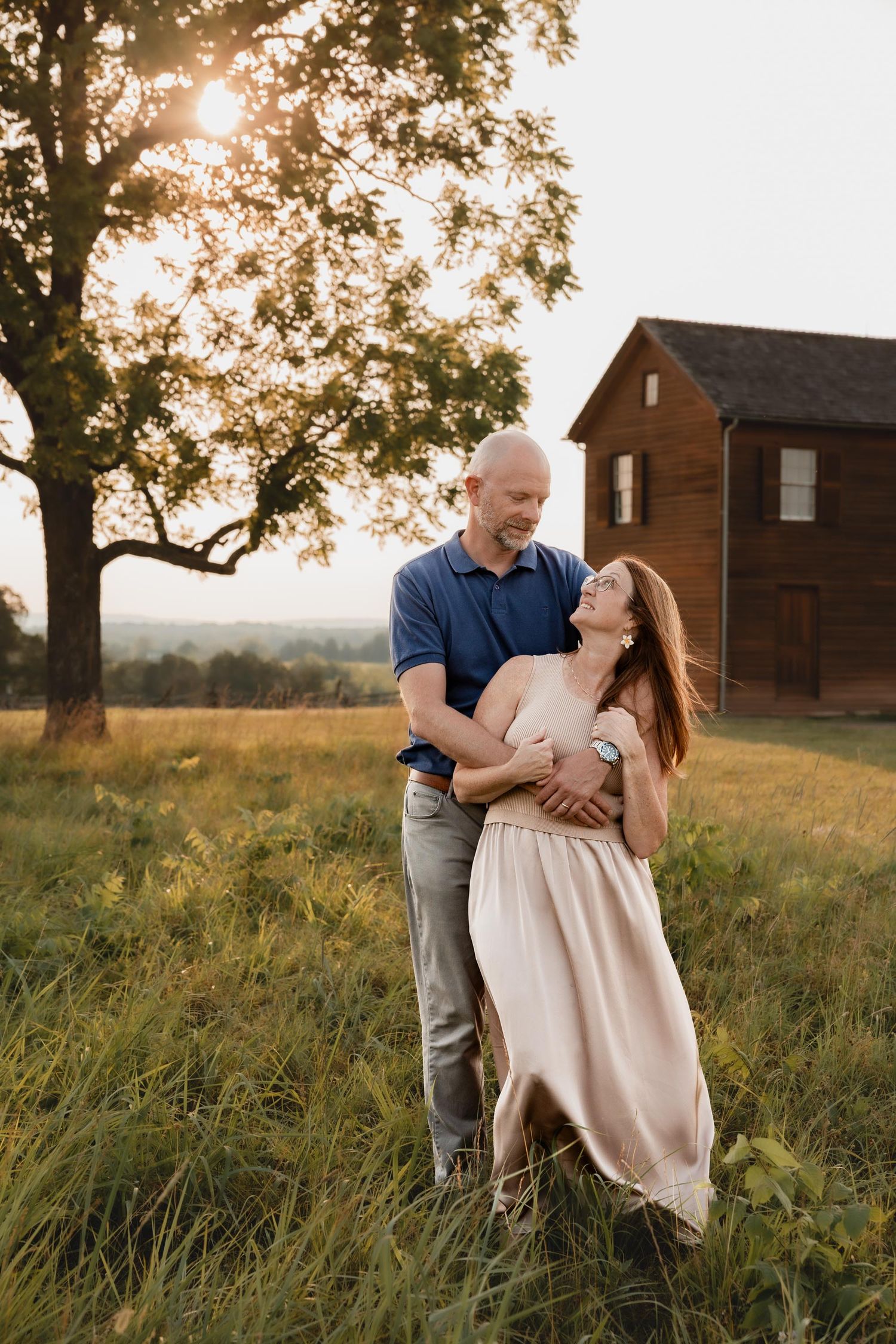A couple embraces in a sunlit field with a large tree and rustic red barn in the background.