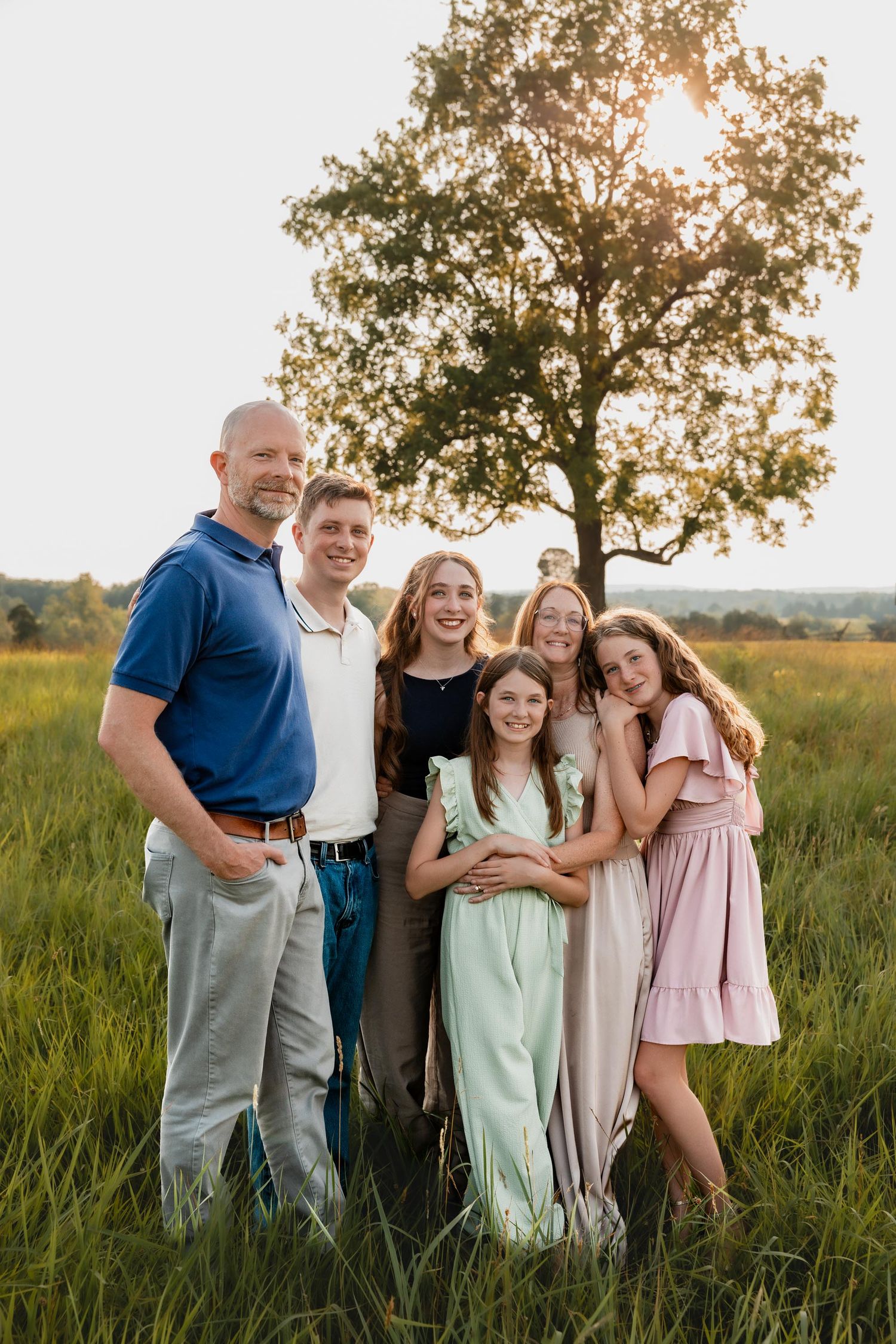 A family of six poses together in a sunlit green field with a large tree backdrop at sunset.