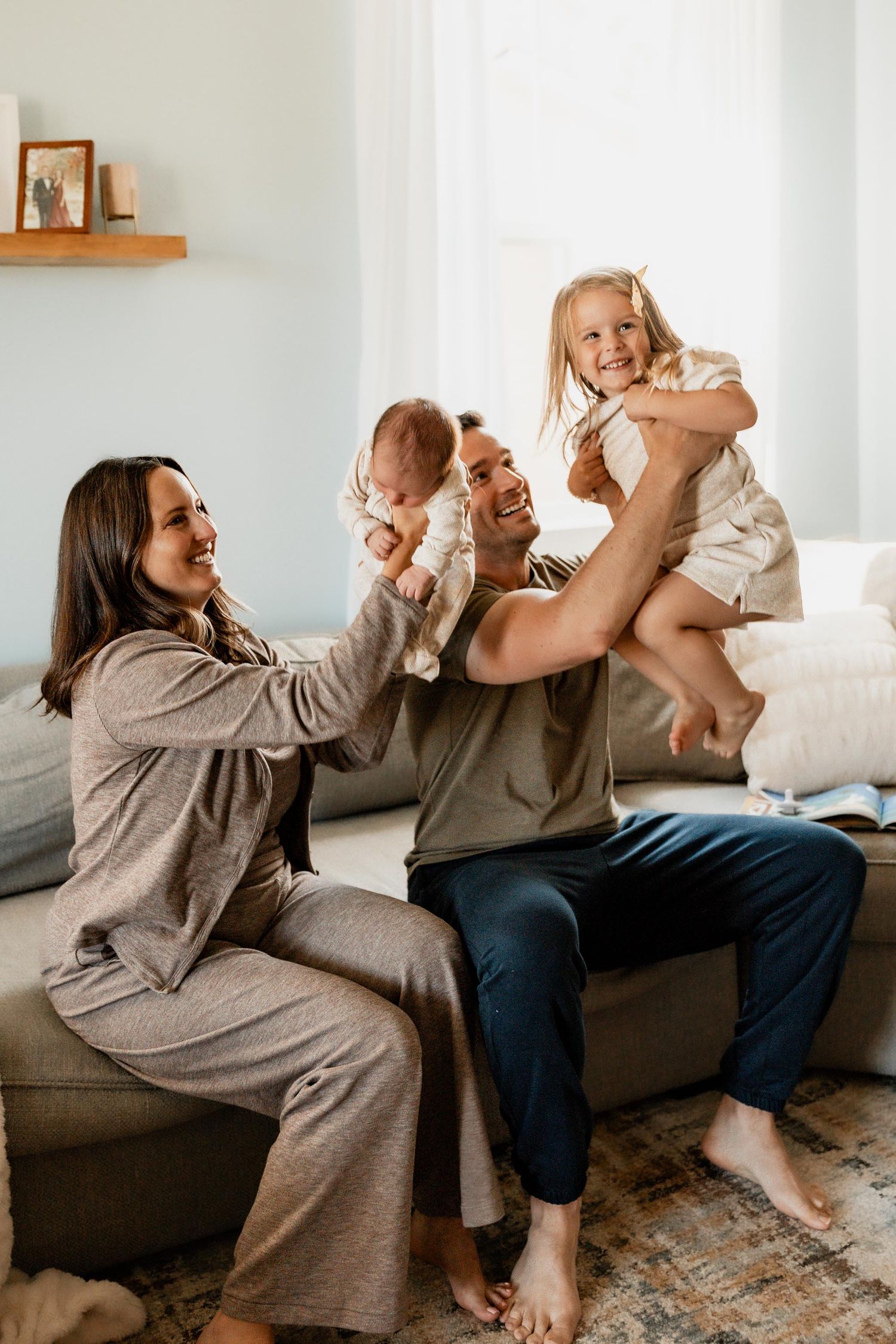 A happy family of four plays together on a couch, with dad lifting toddler girl and newborn baby.