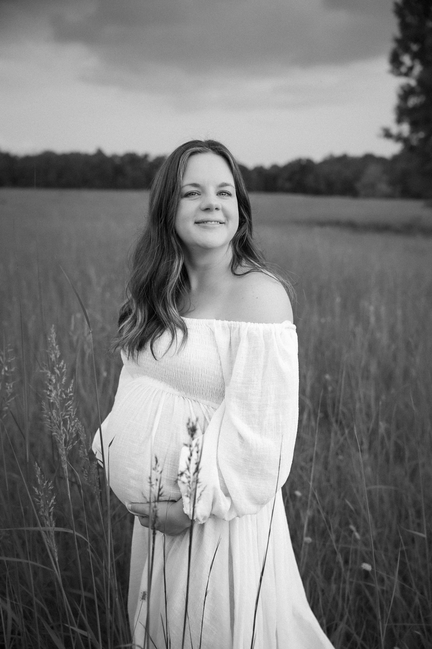 Pregnant woman in white off-shoulder dress standing in field holding wildflowers, black and white maternity portrait.