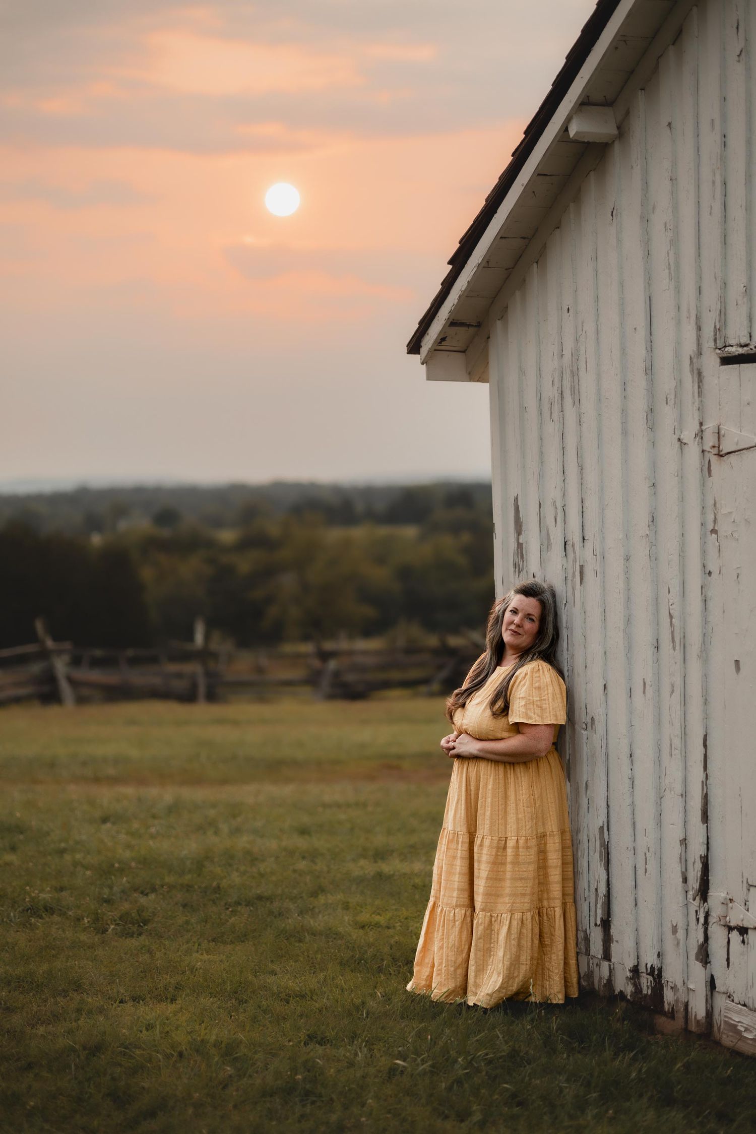 A woman in a yellow maxi dress leans against a white barn wall during a golden sunset.
