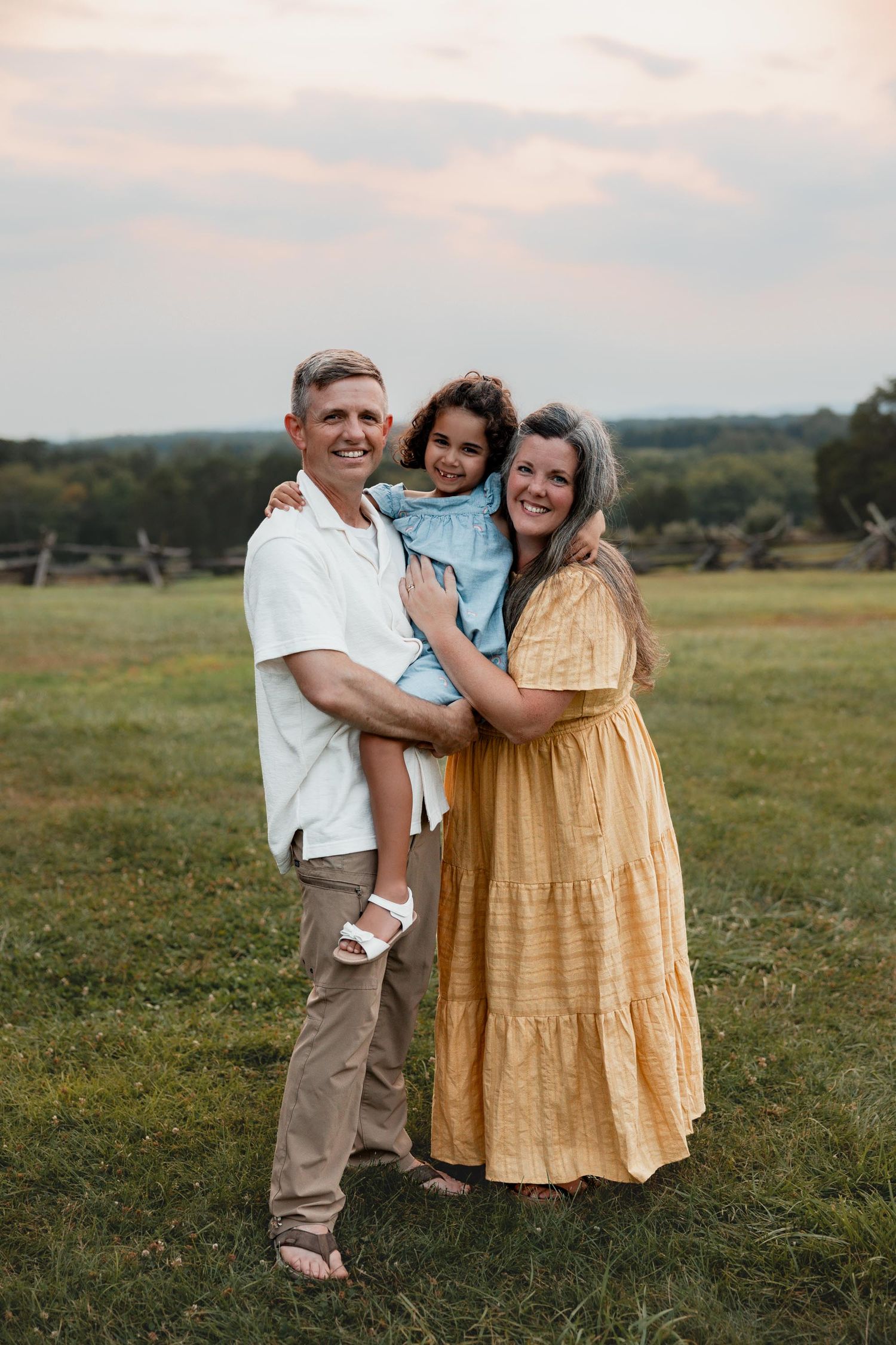 A smiling family of three poses together at sunset in a scenic green field.