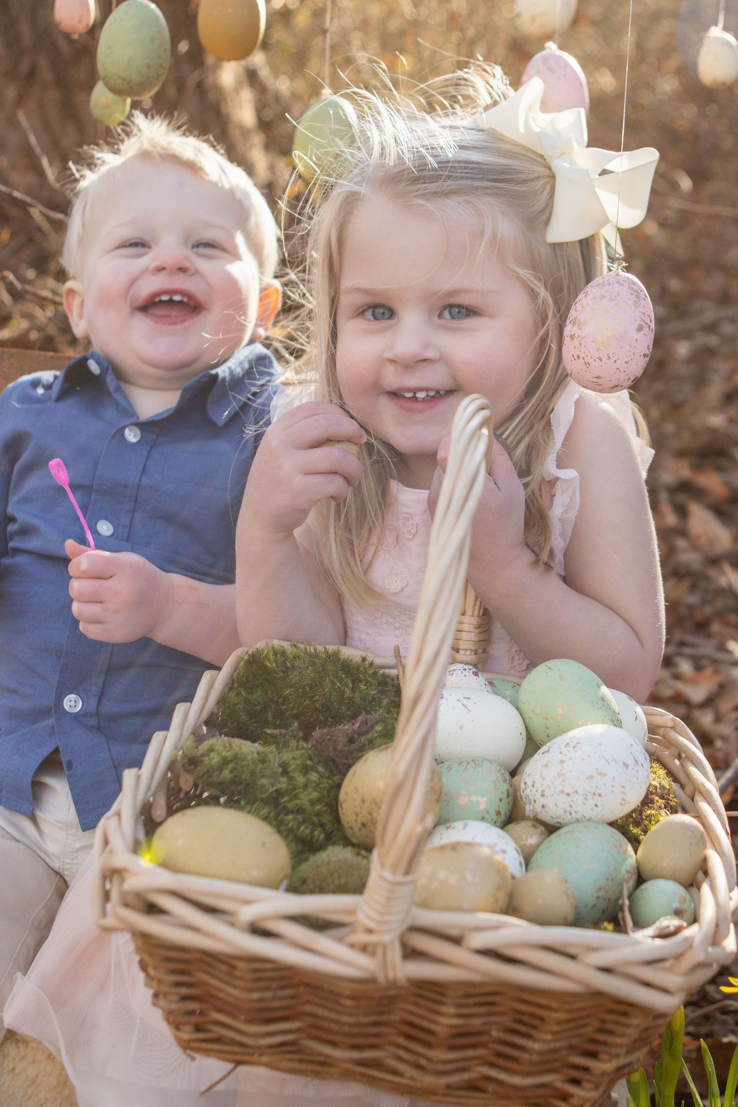 Two young children smile joyfully next to an Easter basket filled with colorful eggs and moss in a springtime setting.