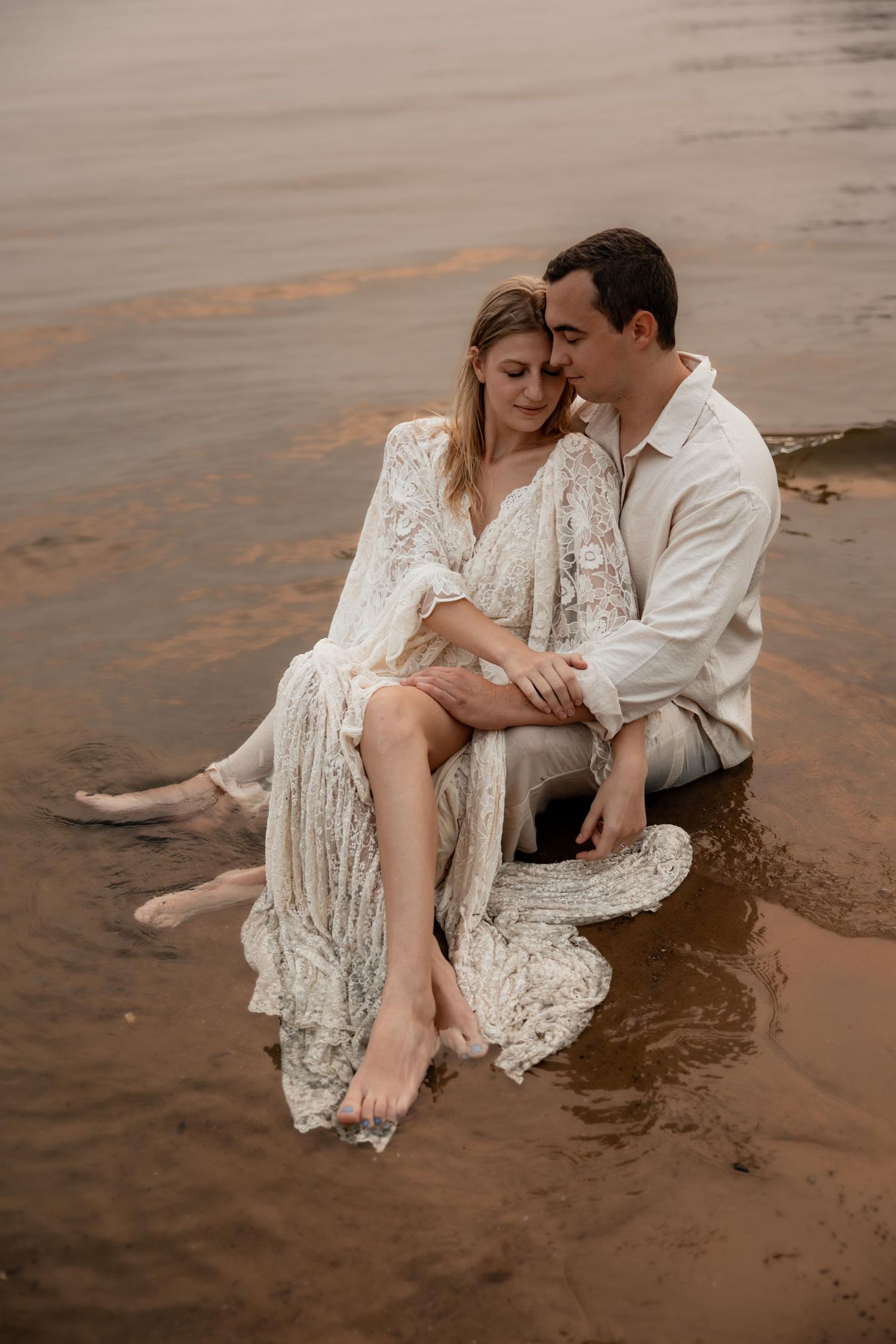 Couple sitting together in shallow water, woman in white lace dress, man in white shirt, romantic beach portrait.