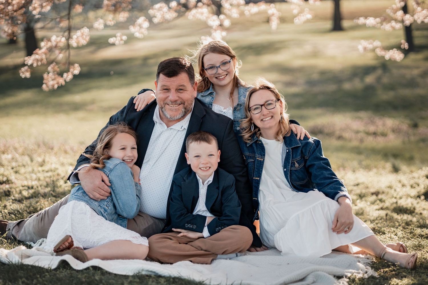 Family of five smiling together on a blanket outdoors, surrounded by blooming cherry trees in soft spring sunlight.