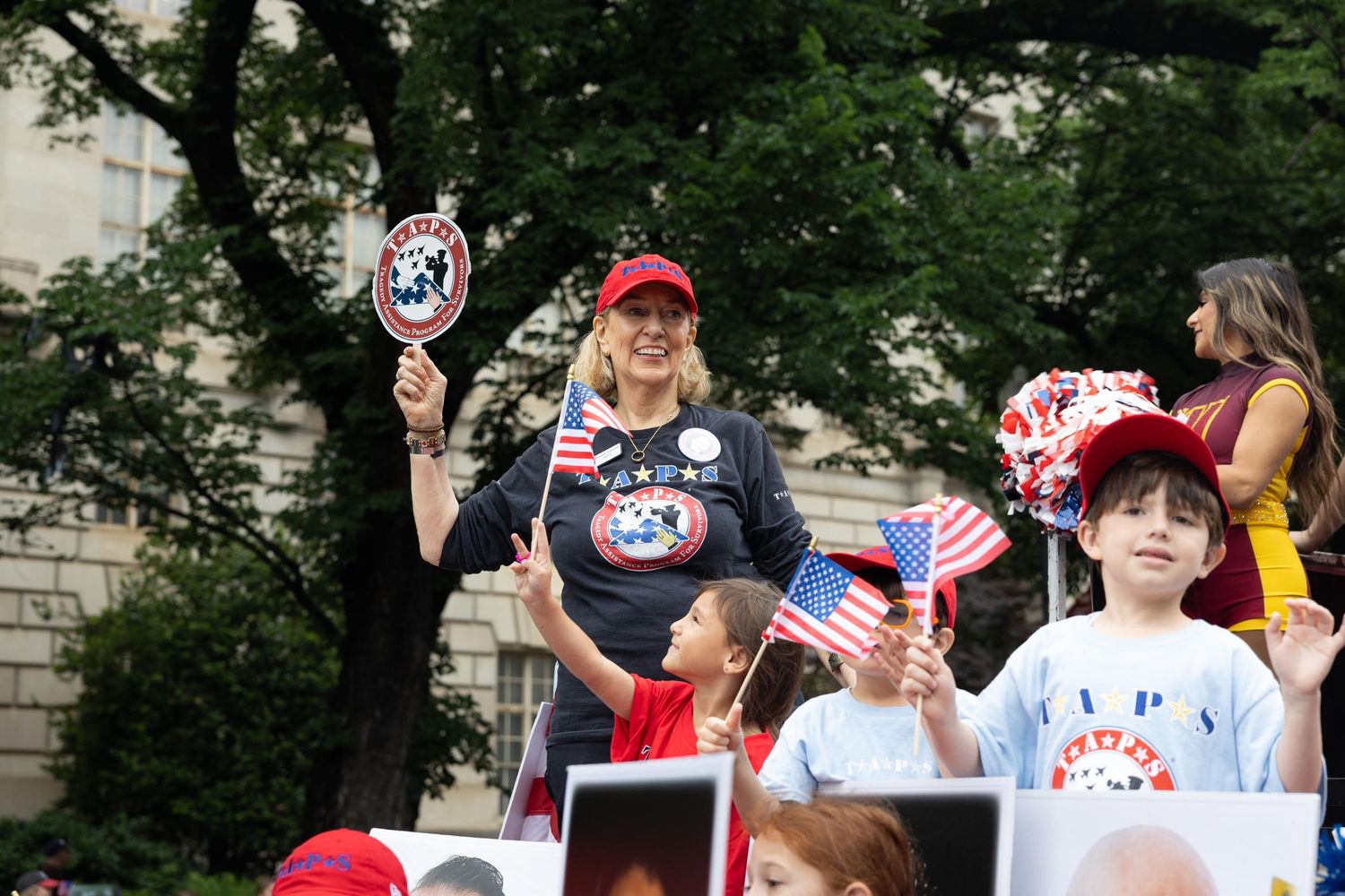 Woman in red cap waves American flags at patriotic rally with children holding flags and signs outdoors.