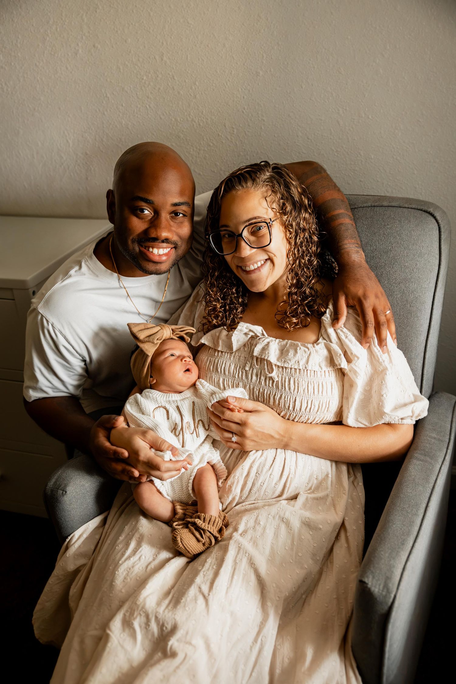 Happy couple sitting in a gray chair smiling while holding their newborn baby dressed in white.