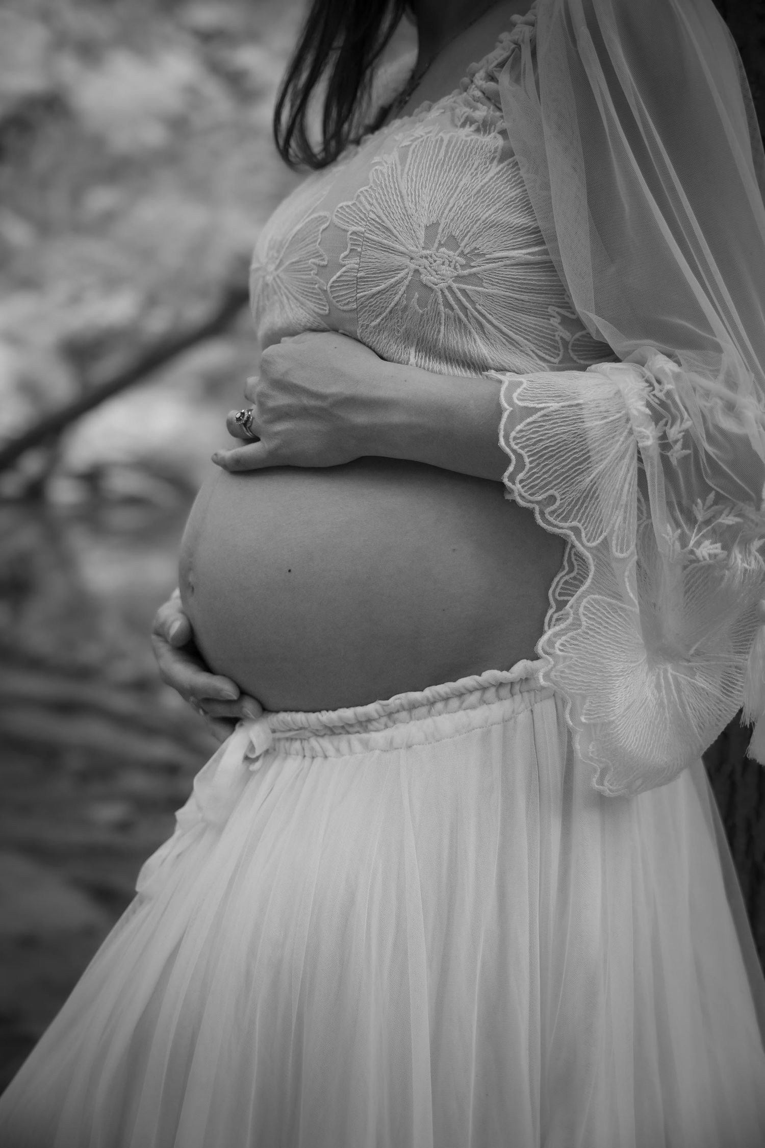 Black and white maternity photo of pregnant woman in lace bridal gown cradling baby bump outdoors.