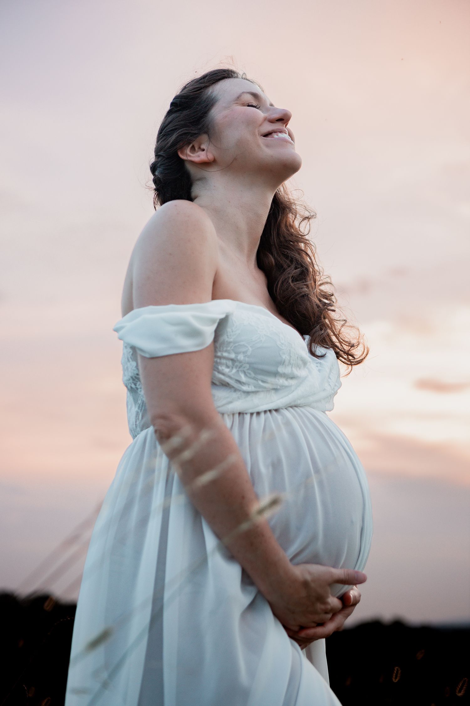 A dreamy maternity portrait in a white flowing dress captures joyful moments at sunset.