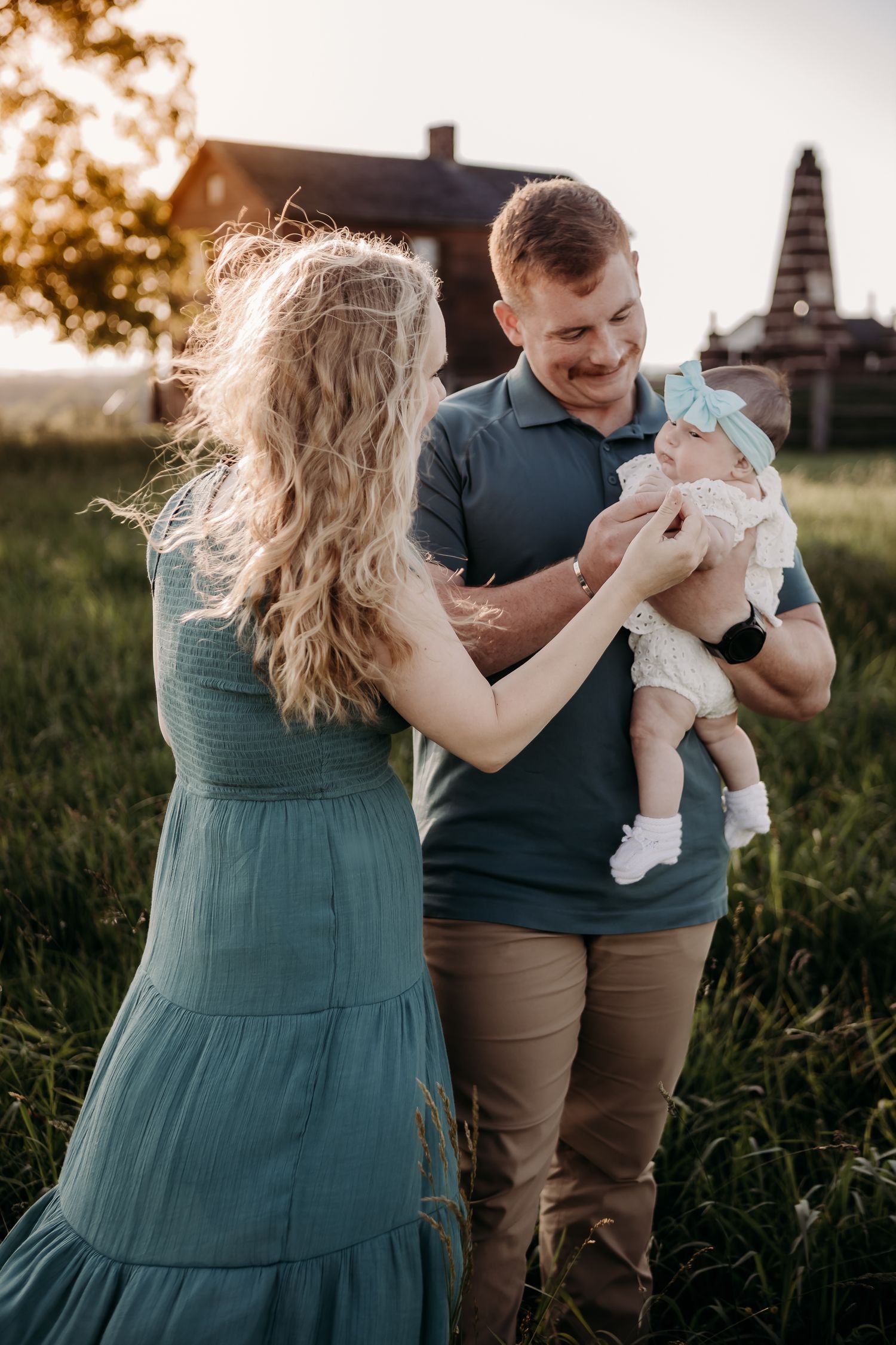 A couple in a teal dress and grey shirt stand together with a baby outdoors at sunset in a grassy field.