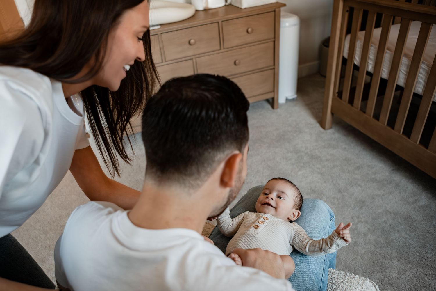 Parents sitting on nursery floor with their smiling infant baby next to a wooden crib and dresser.