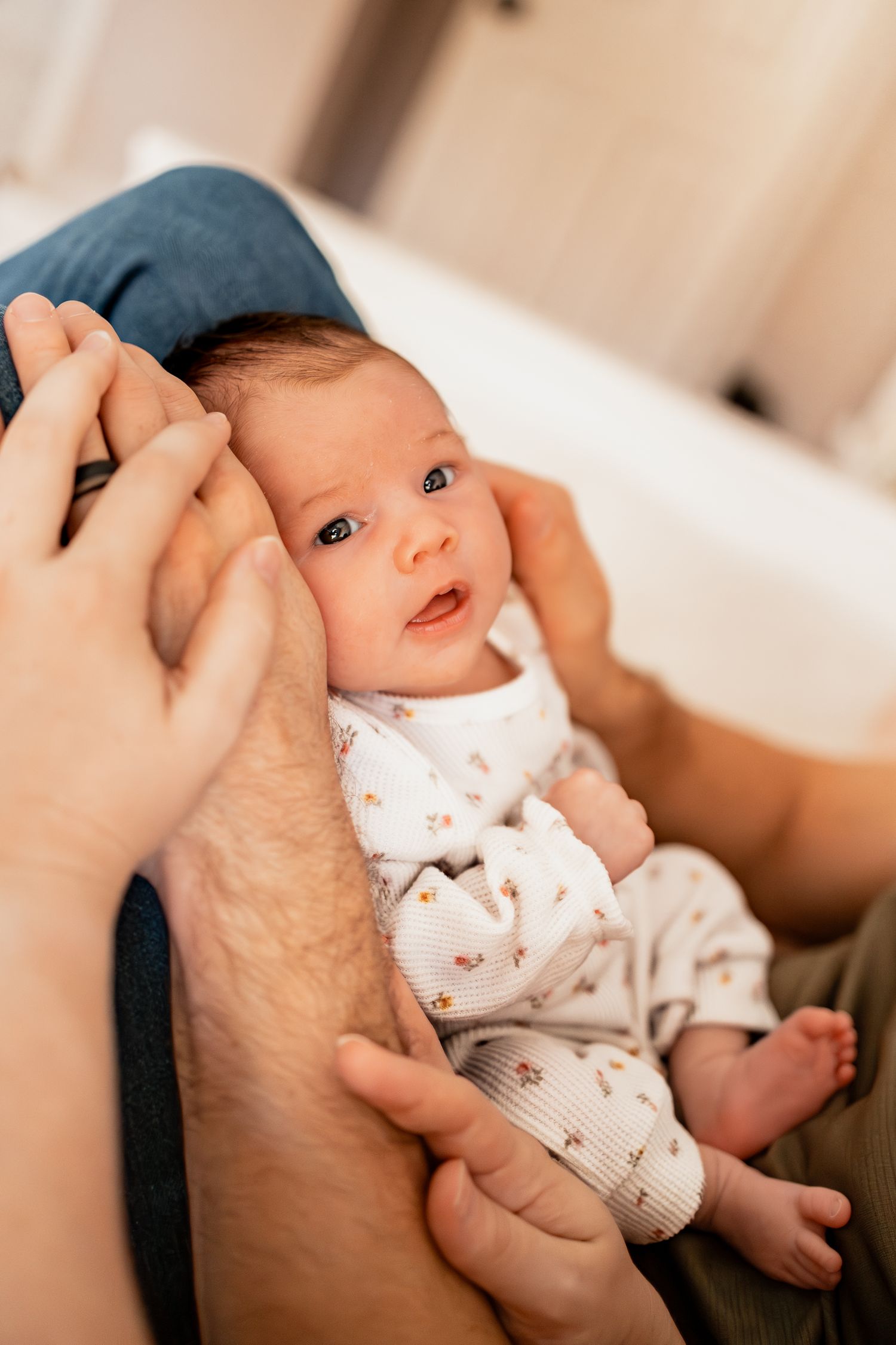 A baby in white clothing is being cradled in someone's arms while looking up with bright eyes.