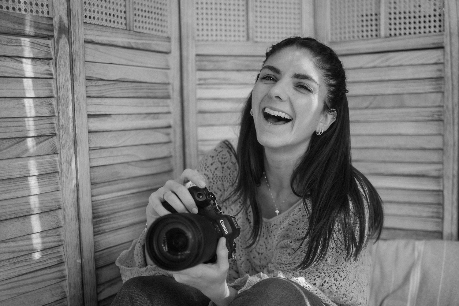 Black and white photo of a smiling woman holding a DSLR camera, sitting in front of wooden shutters.