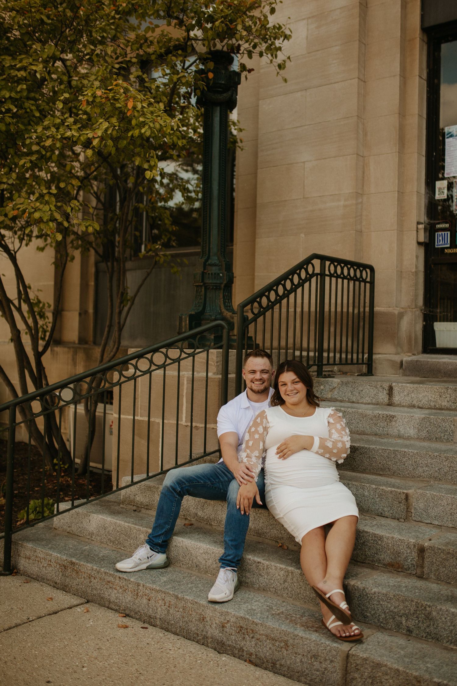 A couple sits together on stone steps outside a building with ornate railings and autumn foliage.