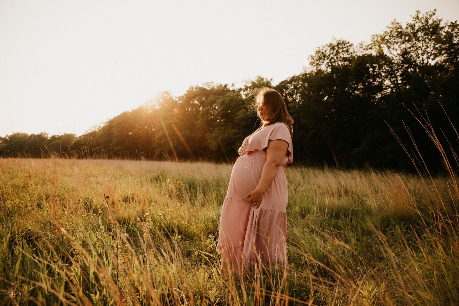 Silhouette of pregnant woman in flowing pink dress standing in sunlit meadow.