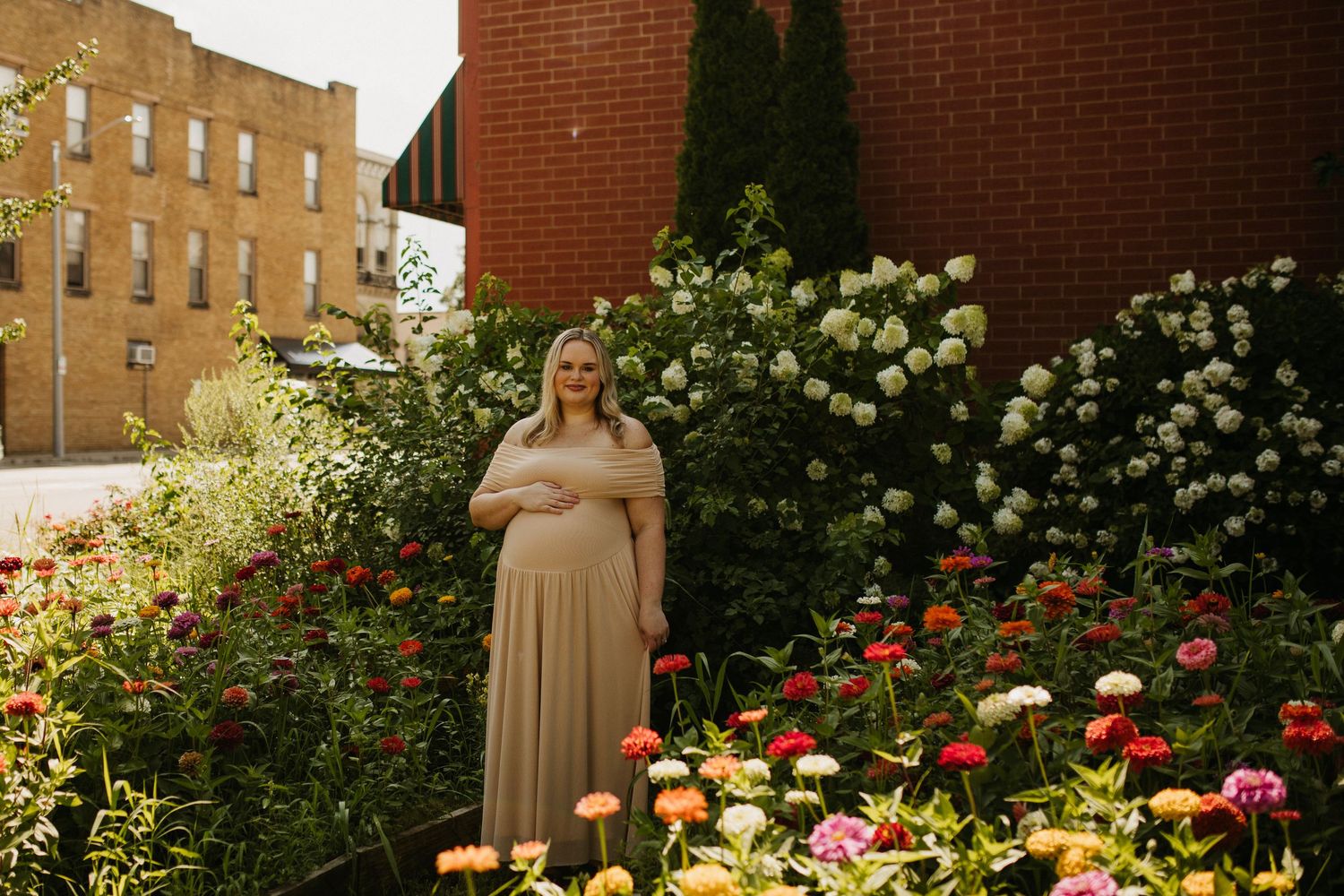 Expectant mother in beige dress poses in garden with colorful flowers and brick building.