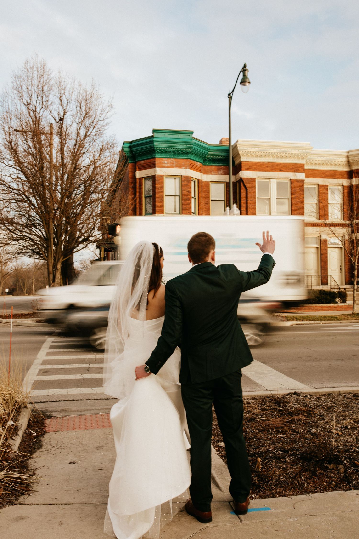 Couple in wedding attire waving while walking down a street with historic brick buildings.