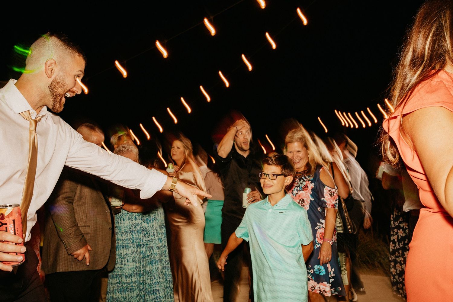Wedding guests dance and celebrate on a dimly lit dance floor with string lights overhead.