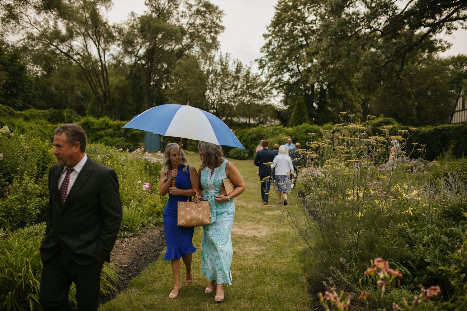 Wedding party walks through a lush garden path holding blue umbrellas on an overcast day.