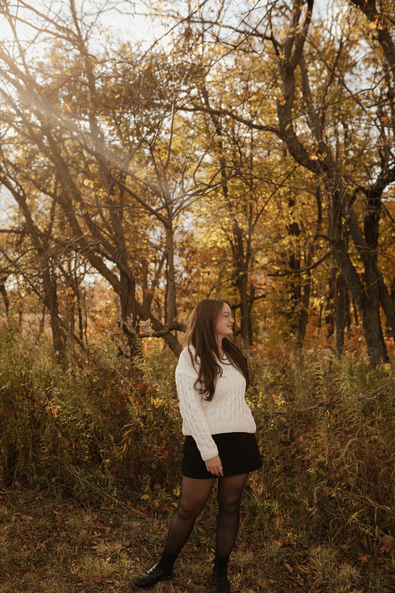 A figure in a white sweater and dark bottoms poses against autumn foliage in a wooded outdoor setting.