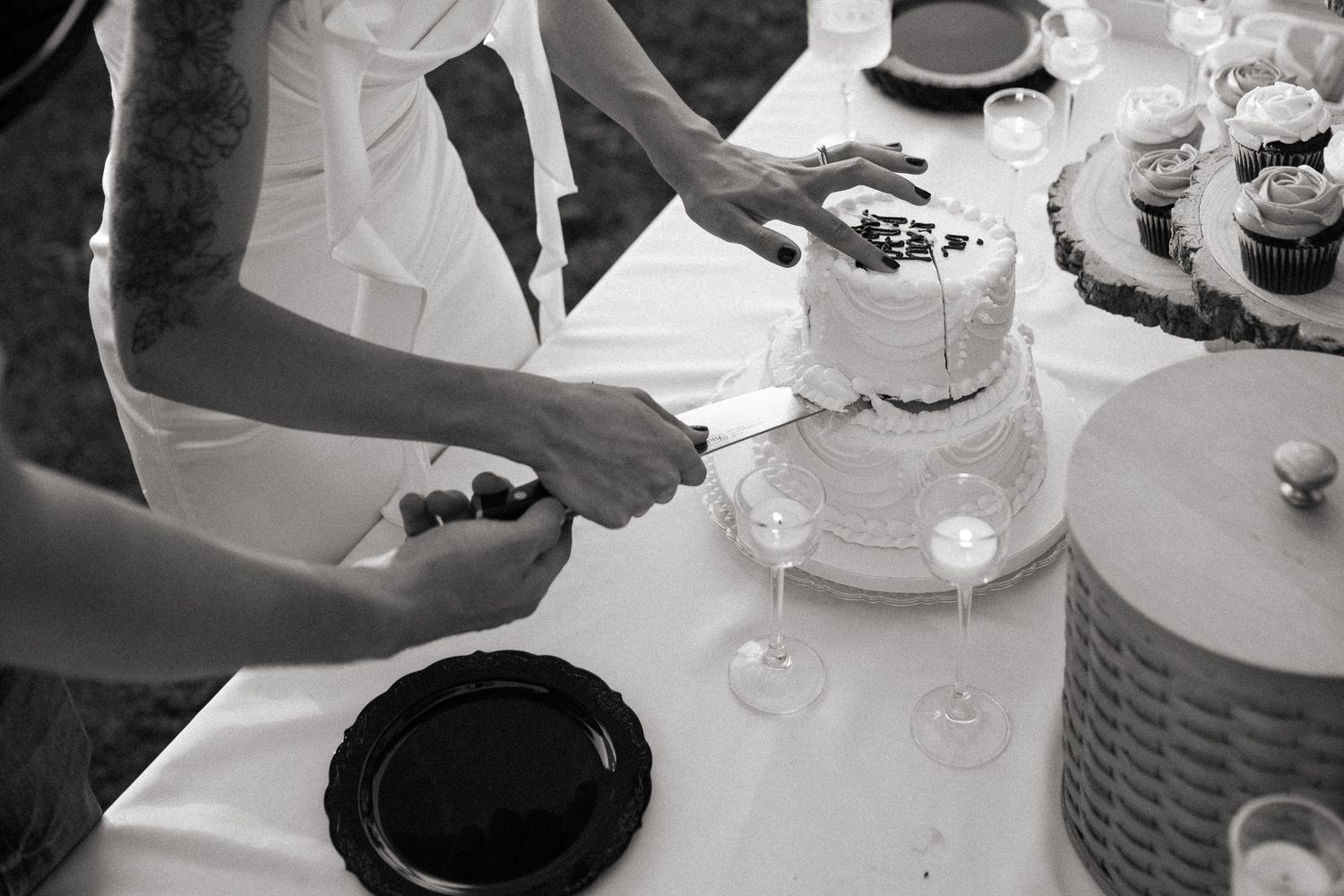 Black and white photo of hands cutting a wedding cake with plates and glasses on a table.