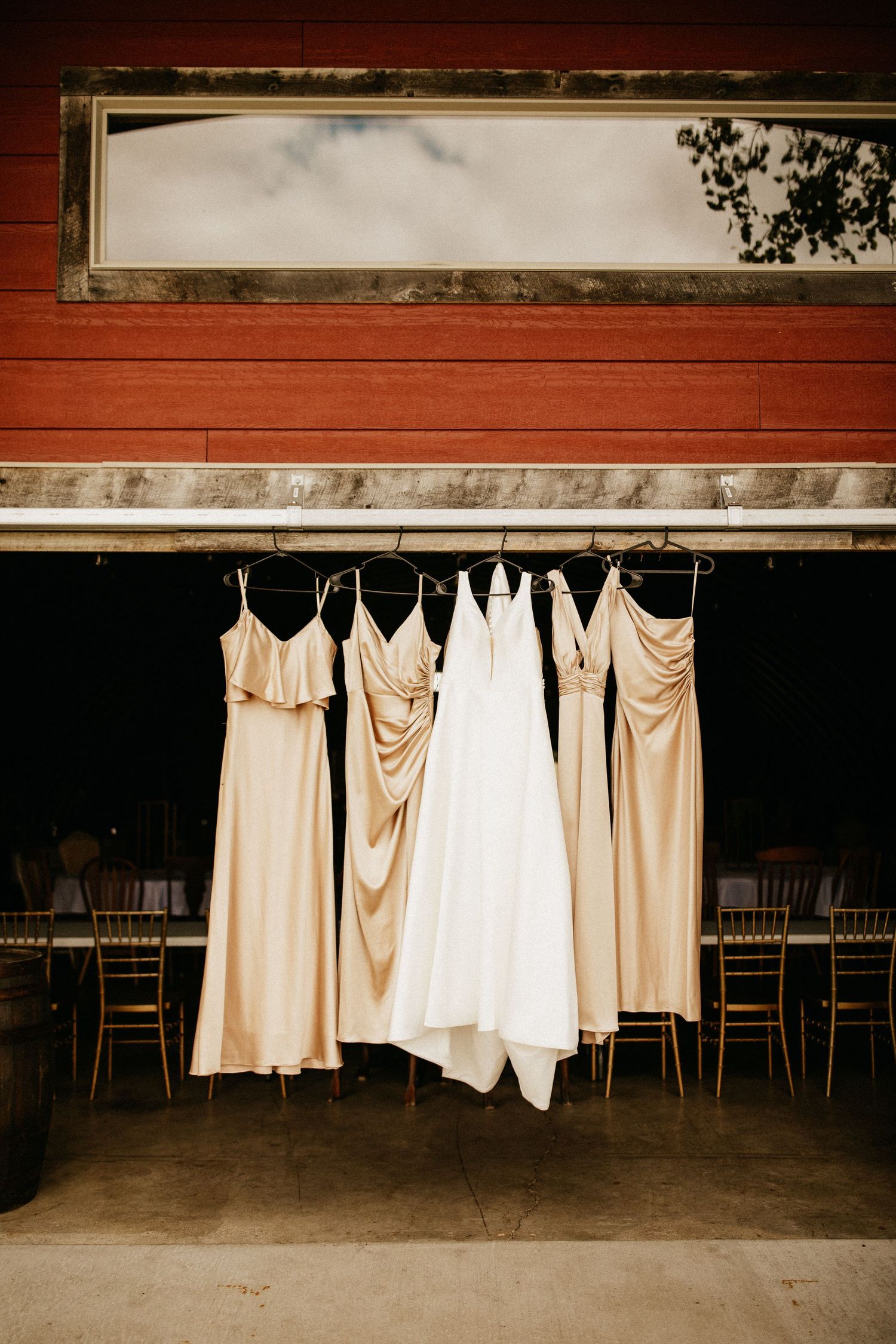 Row of light colored bridesmaid dresses hanging against a dark wooden wall.
