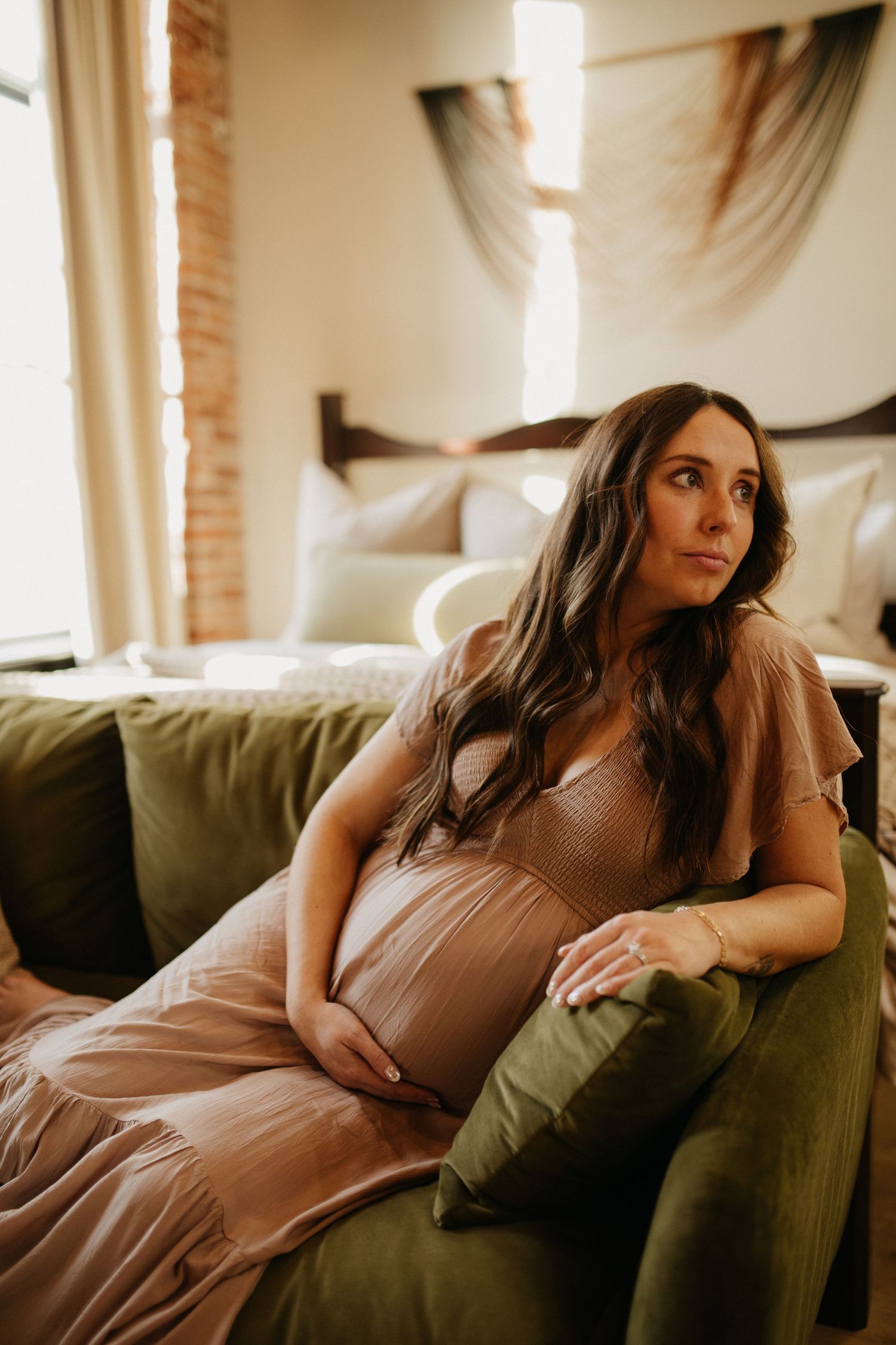 Expectant mother in pink dress rests on olive green couch near window with brick wall.