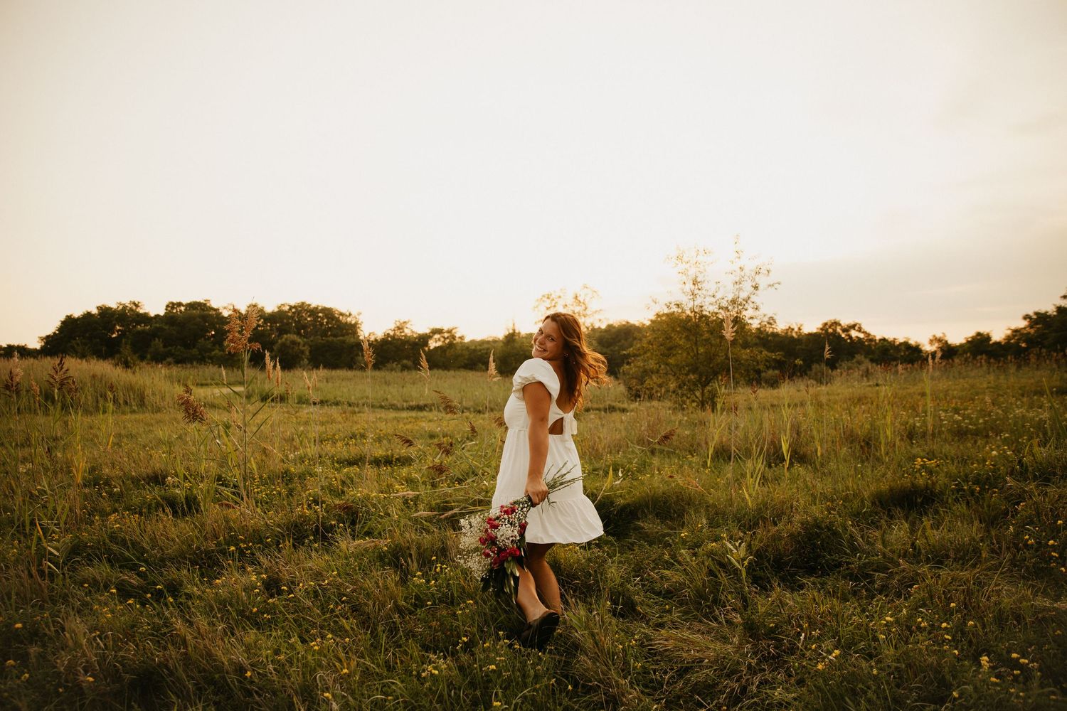 Person in white dress walks through tall grass field at golden hour.