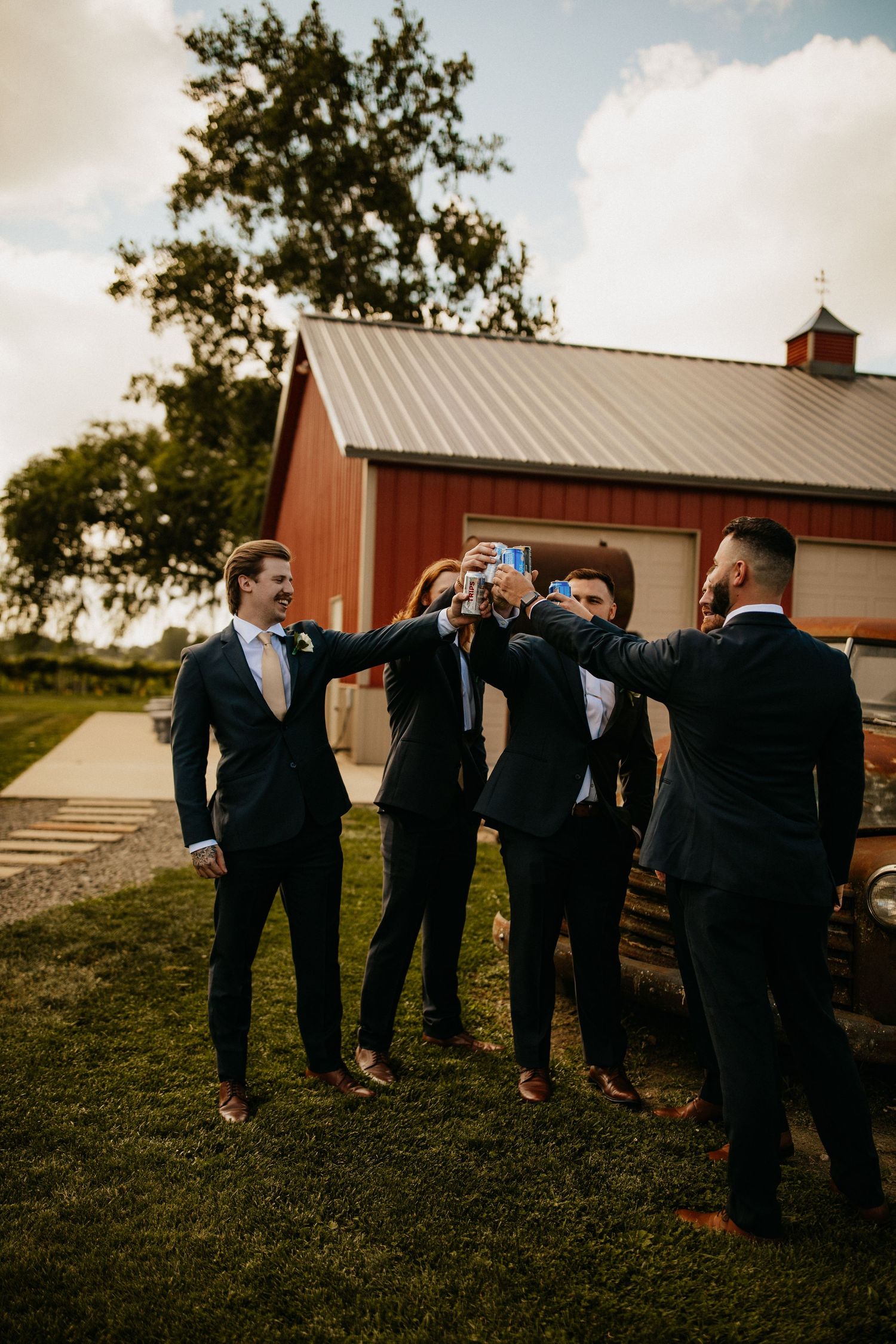 Groomsmen in black suits pose playfully on a grassy hill near a red barn.