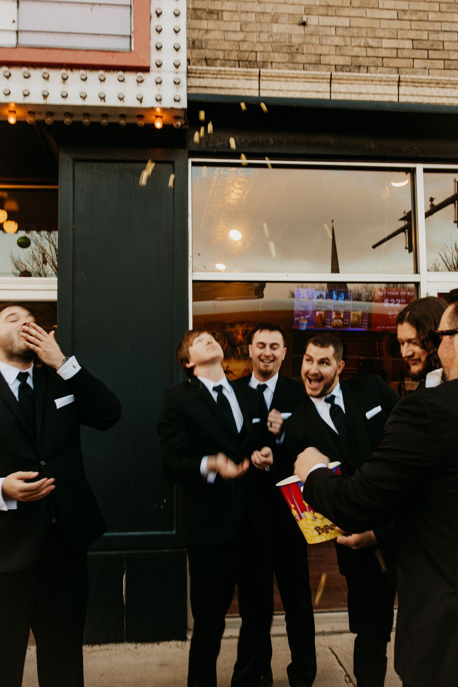 Group of groomsmen in black suits laughing and celebrating in a dimly lit venue.