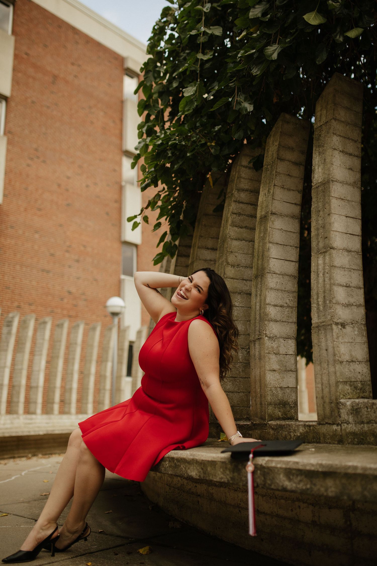 Woman in red dress poses playfully against stone columns in warm sunlight.
