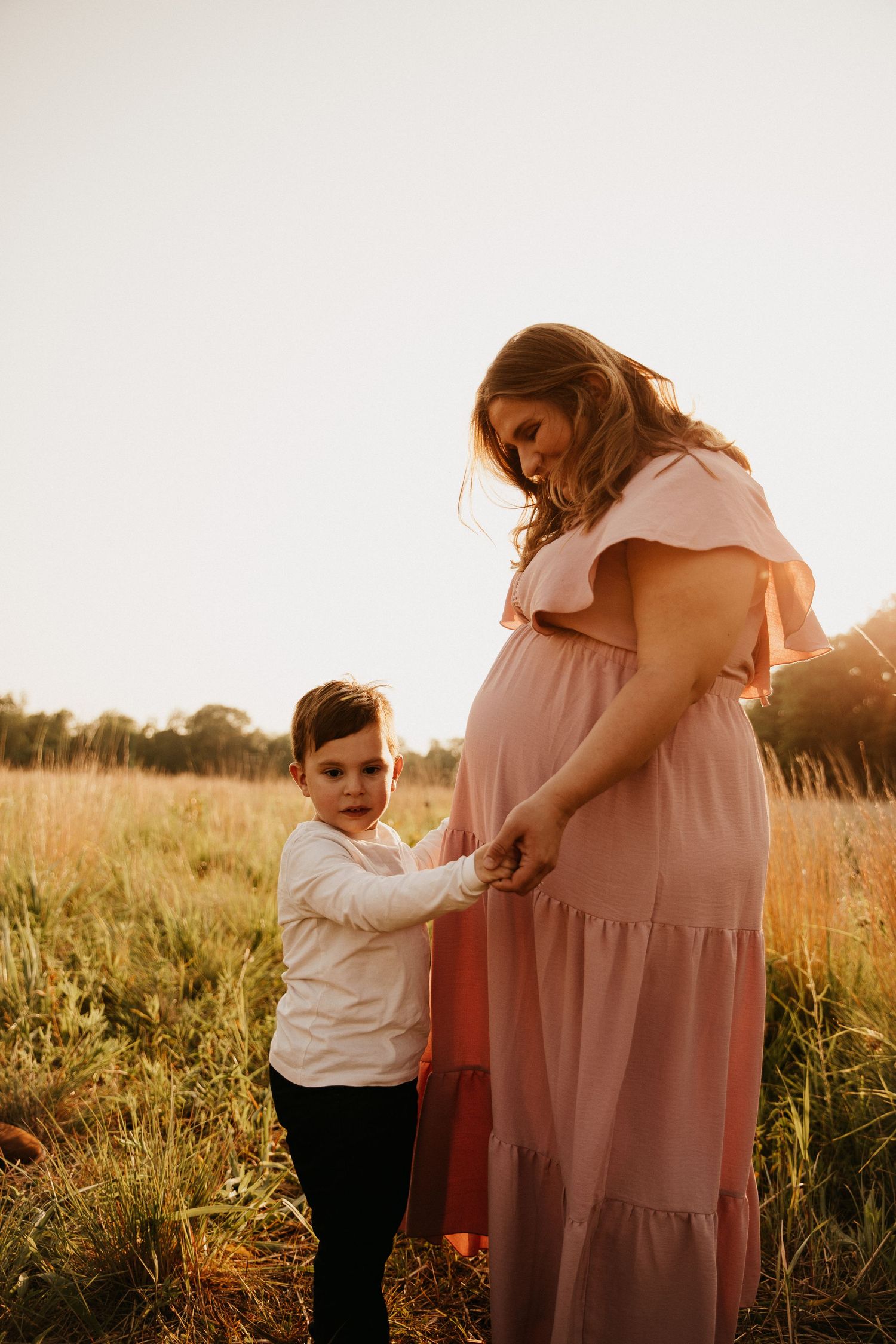 Maternity moment captured in field at sunset with child touching mother's dress.