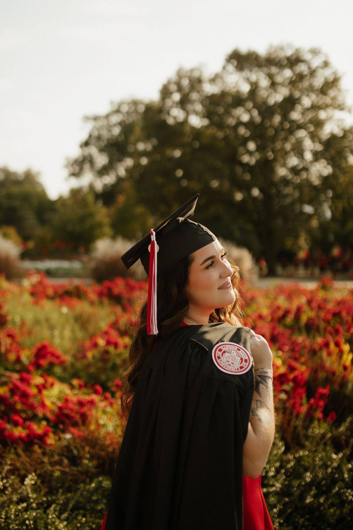 A graduate in black cap and gown poses in a garden with red flowers at sunset.