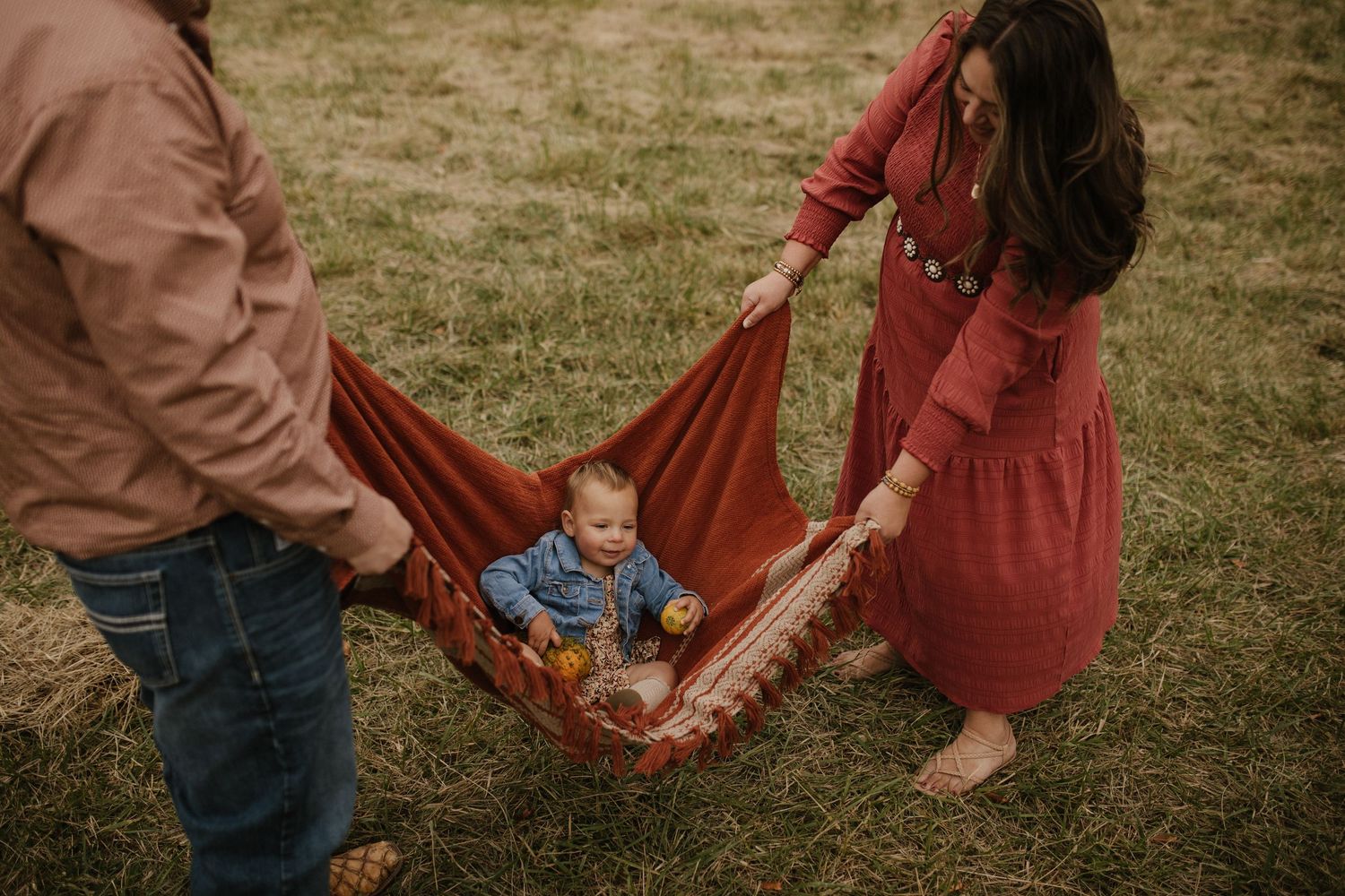 Parents swing a small child in a blanket hammock outdoors on a grassy field.