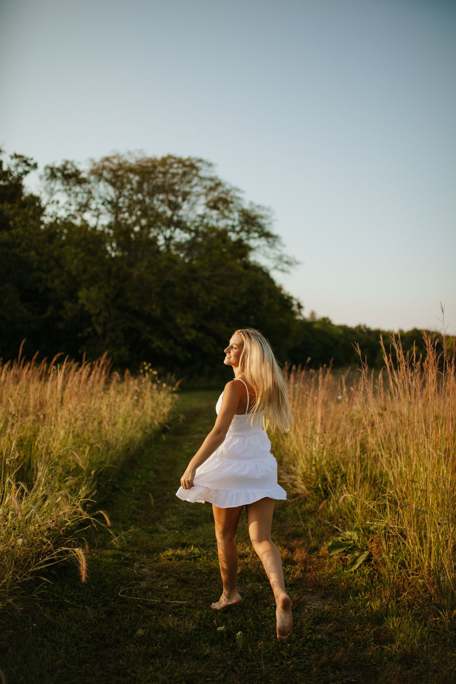 Figure in white dress dances through tall grass at sunset.