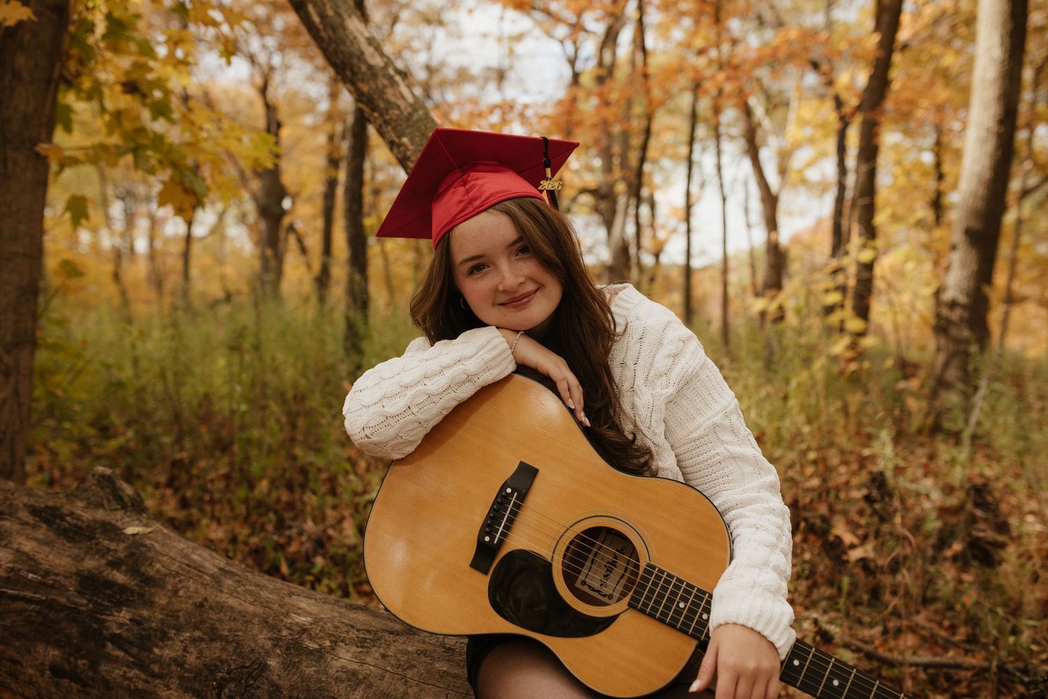 A guitarist in white sweater performs on acoustic guitar in a wooded autumn setting.