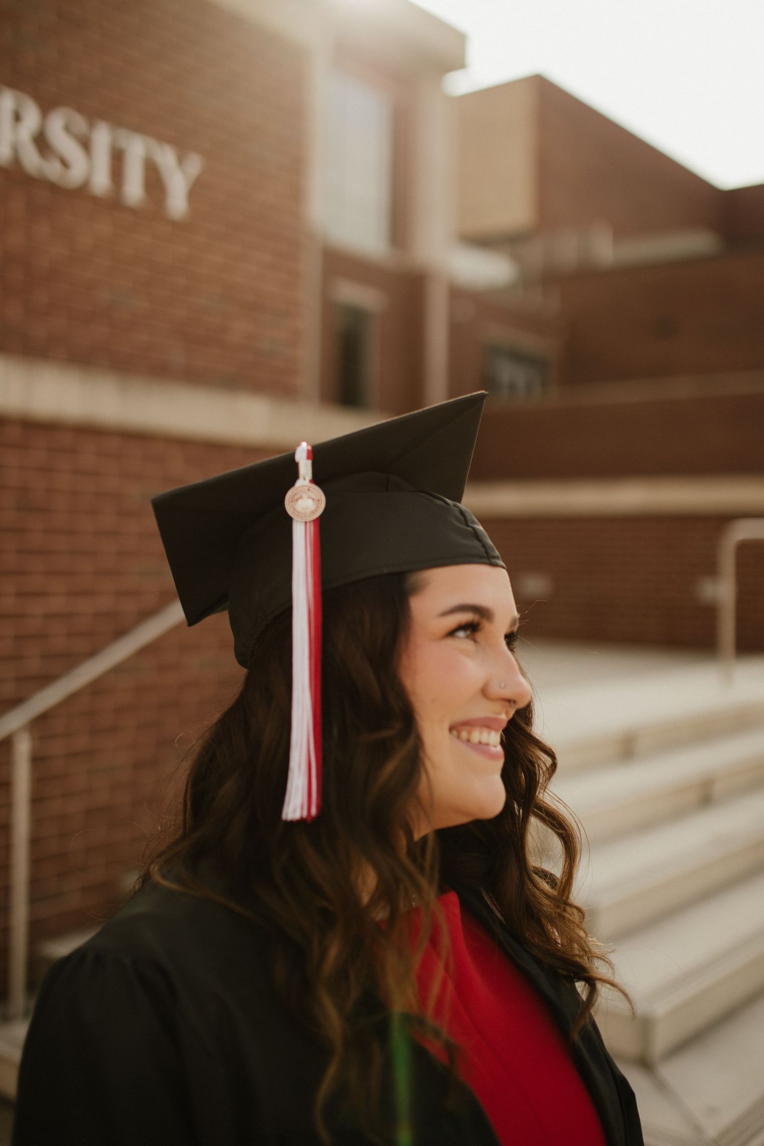 A graduate in cap and gown smiles while sitting on brick steps of a university building.