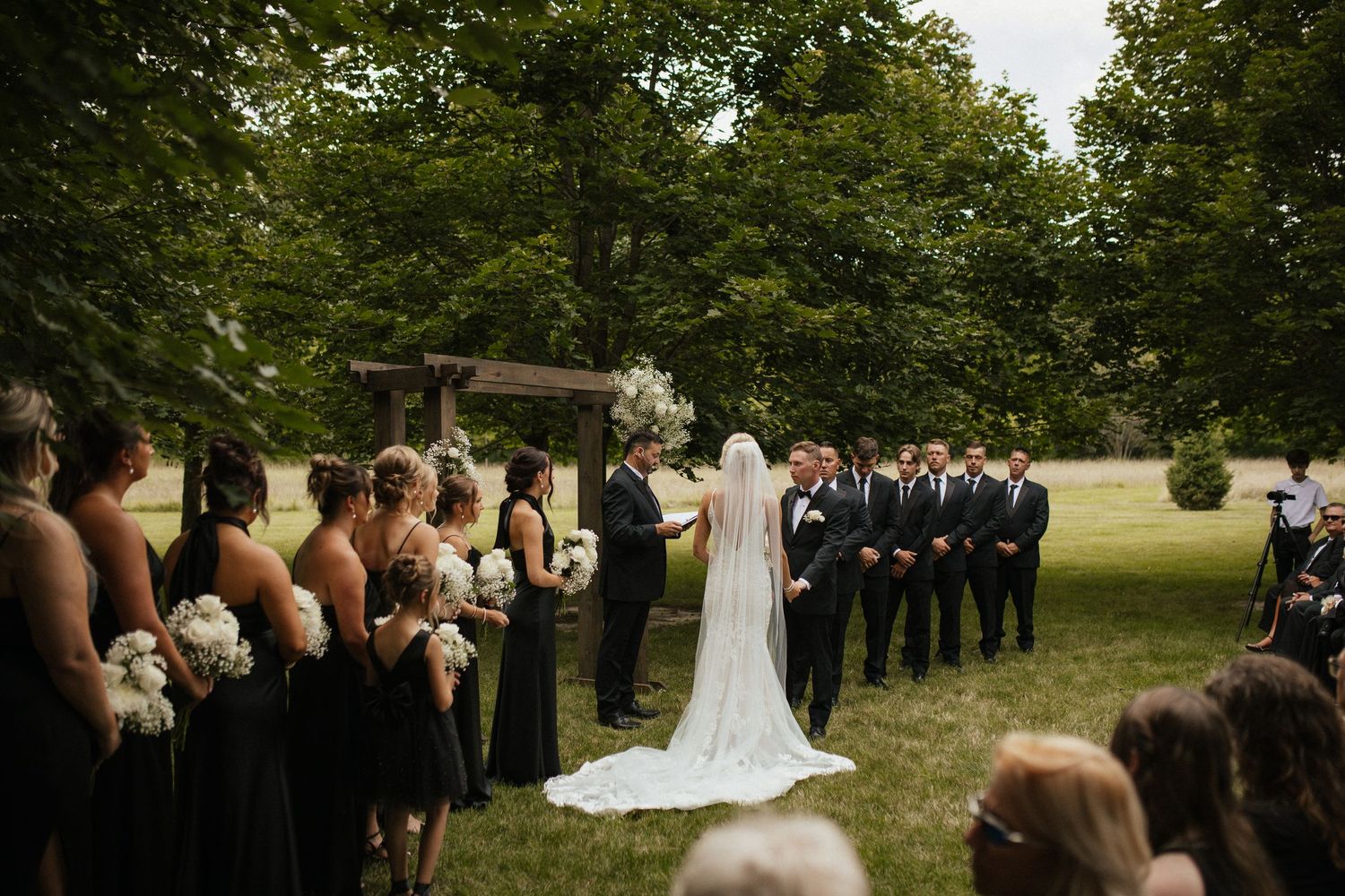 Wedding ceremony guests seated in white chairs on a grassy lawn surrounded by trees.