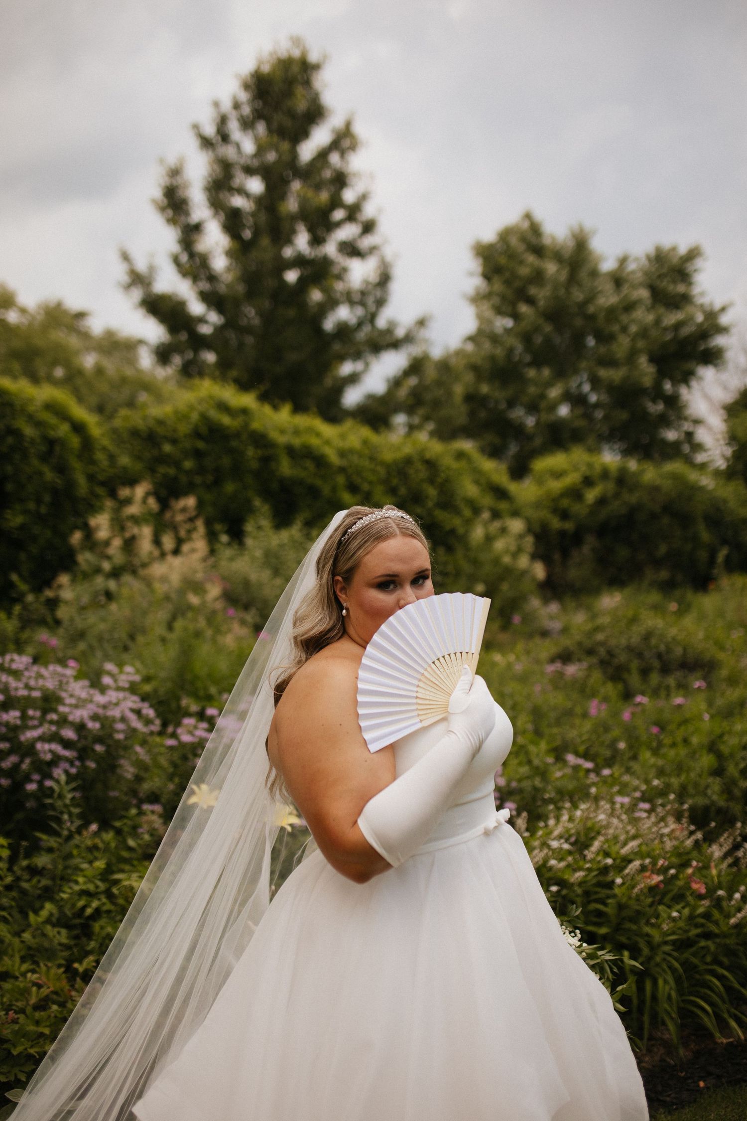 Bride in flowing white dress poses with decorative fan in garden setting.