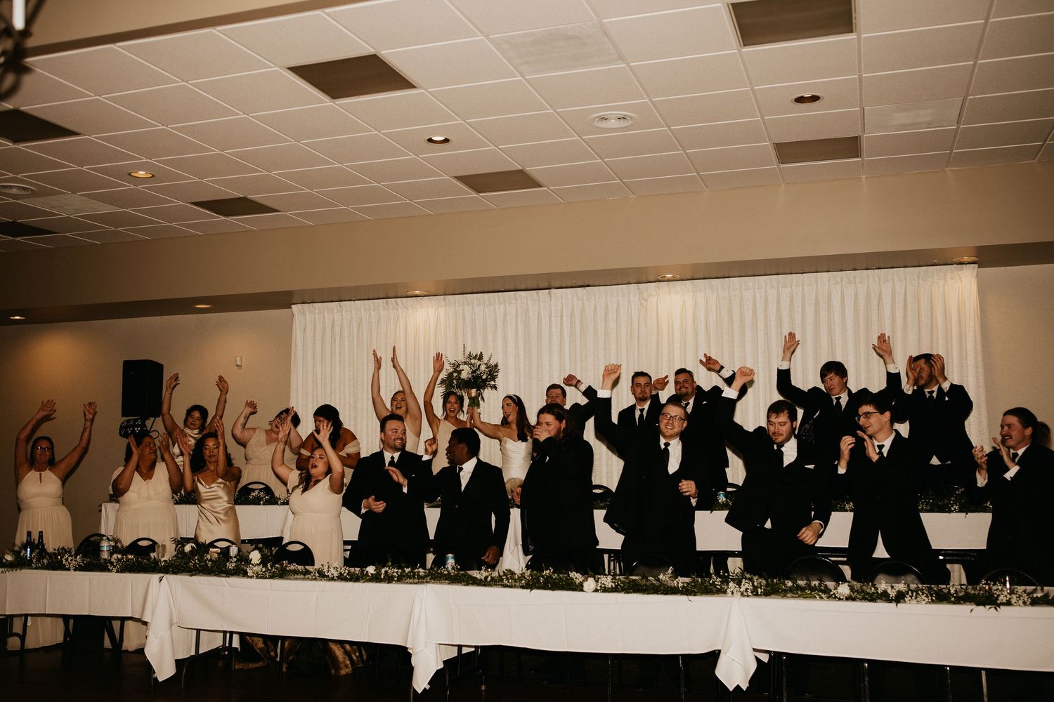 Wedding party members celebrating with raised arms at a reception head table.